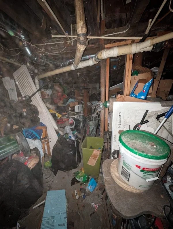 Cluttered basement storage area with pipes, boxes, buckets, and various tools and debris scattered around.