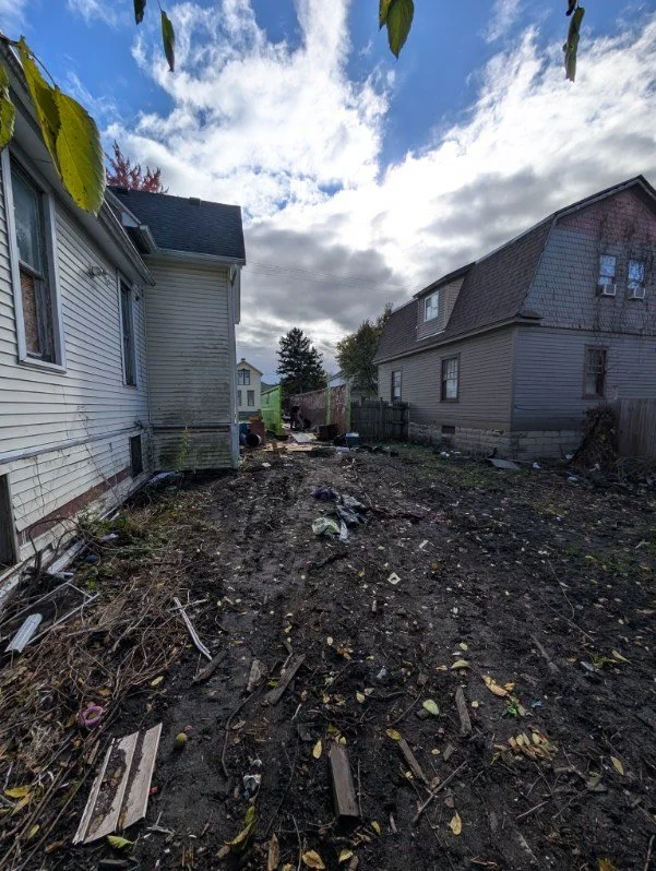 Backyard with dirt and scattered trash, between two houses with cloudy sky overhead.