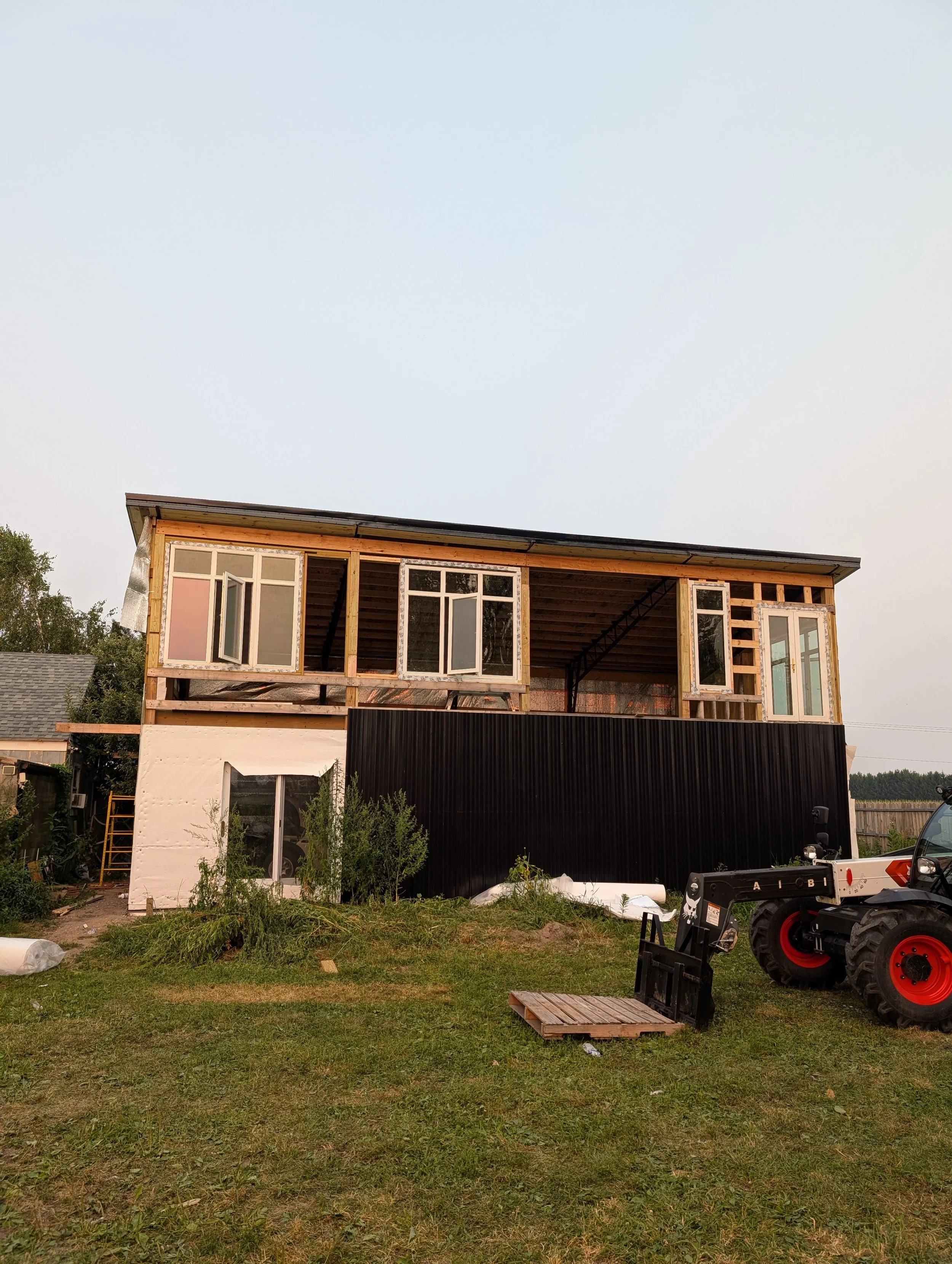 Two-story house under construction with open windows, black exterior wall on the lower level, and construction equipment on a grassy yard.