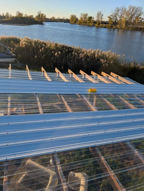 Partially constructed greenhouse with metal framing and transparent panels overlooking a river with trees in the background.