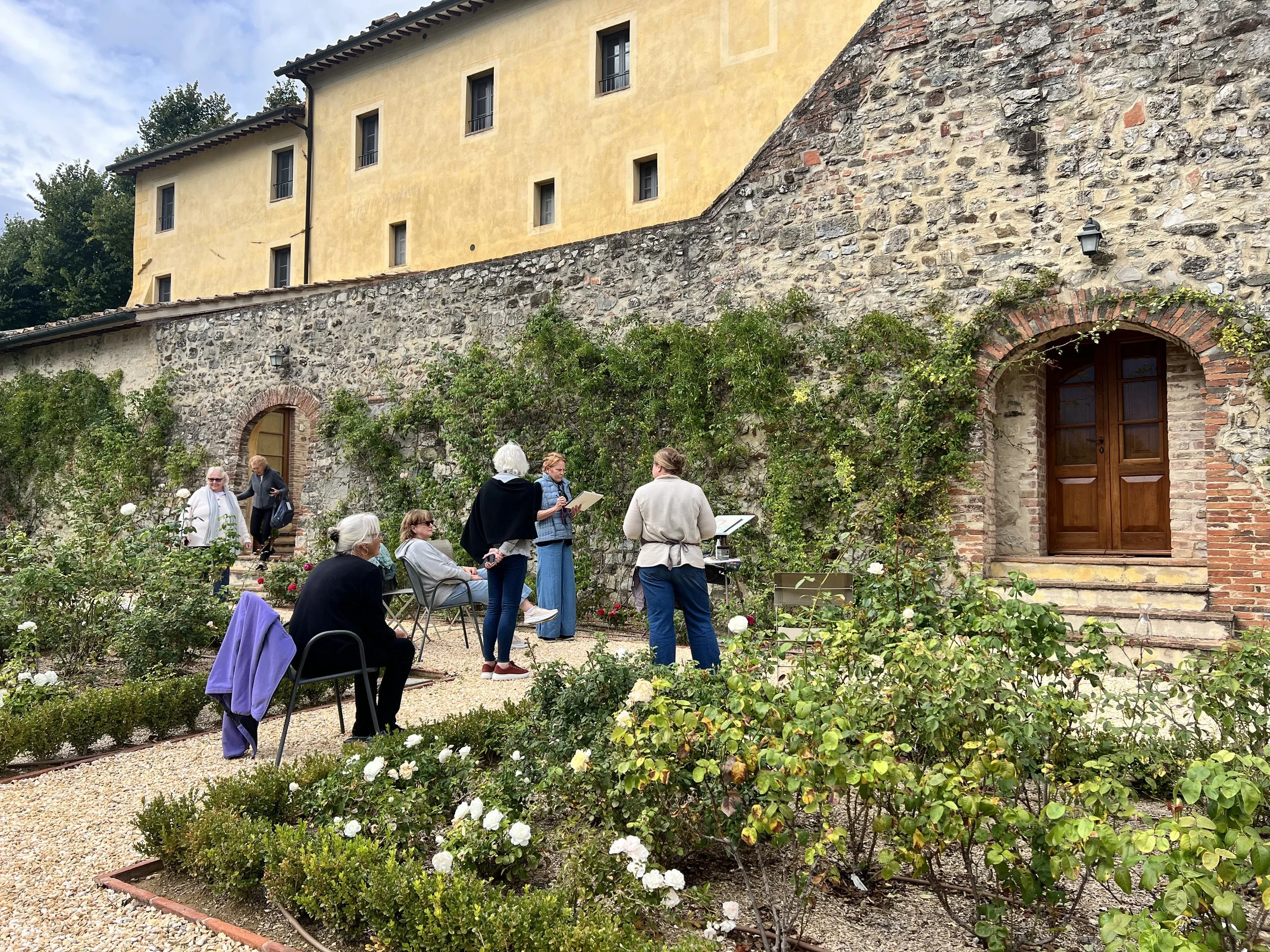 A group of people gathered outside a historic stone and brick building with yellow walls, some sitting and some standing, engaging in a discussion or tour, surrounded by a garden with white flowers.