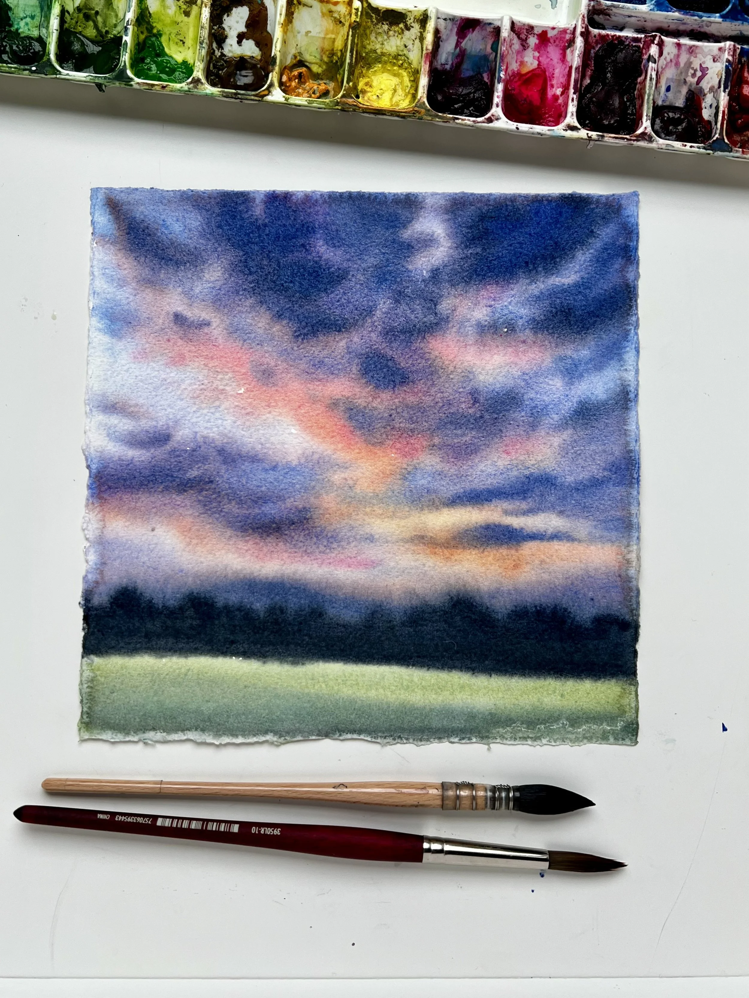 A small watercolor painting of a sunset or sunrise sky with dark clouds, pink, orange, and purple hues, over a green landscape. Two paintbrushes are placed below the painting on a white surface, with an open watercolor palette visible at the top of the image.