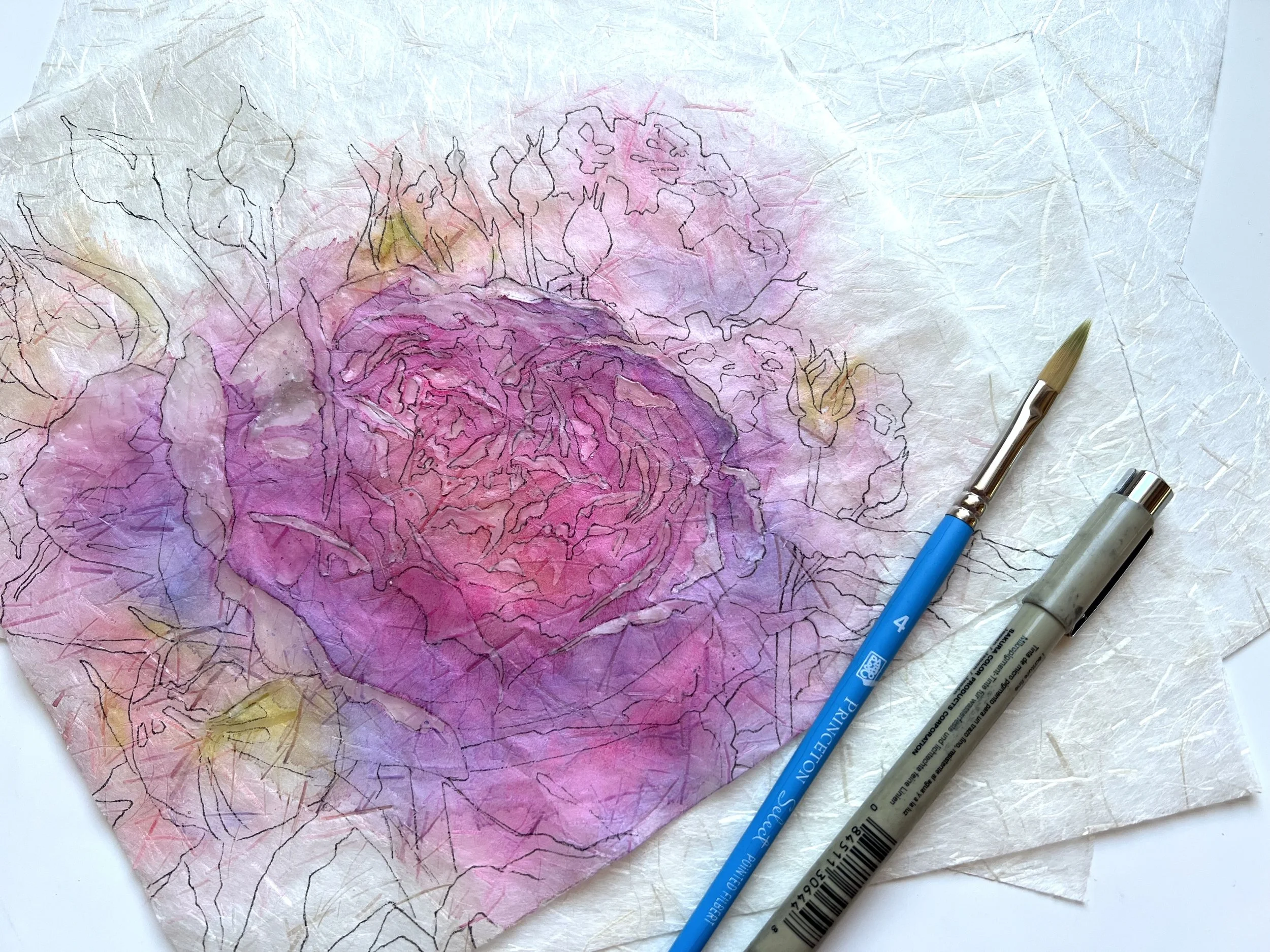 A watercolor painting of a large pink and purple flower, possibly a peony, on textured handmade paper. Two paintbrushes, one with a blue handle and one with a gray handle, are placed on the paper.