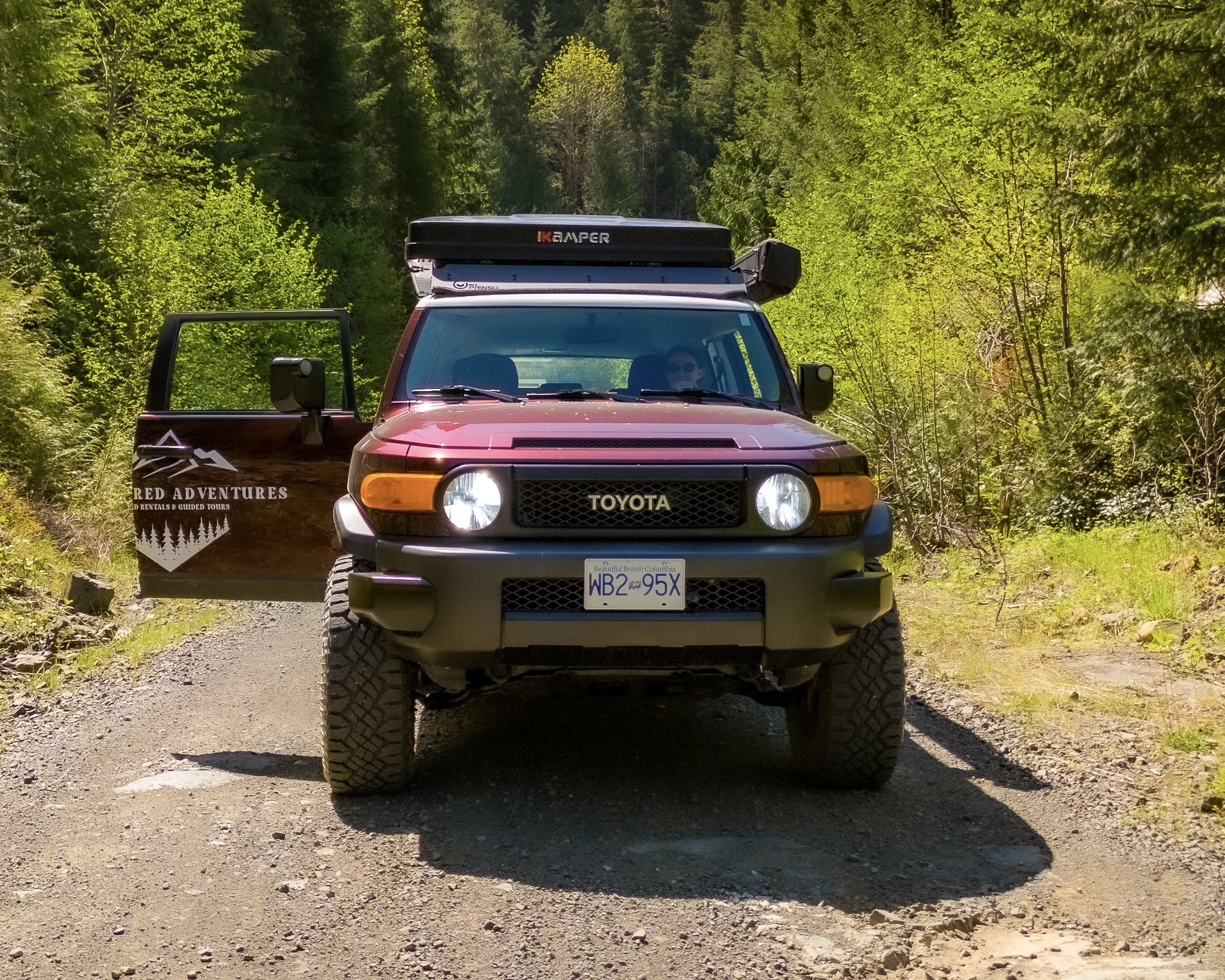 A red off-road vehicle with gear on top and a spare tire at the back parked on a rocky beach, with green forested hills and mountains in the background under a cloudy sky.