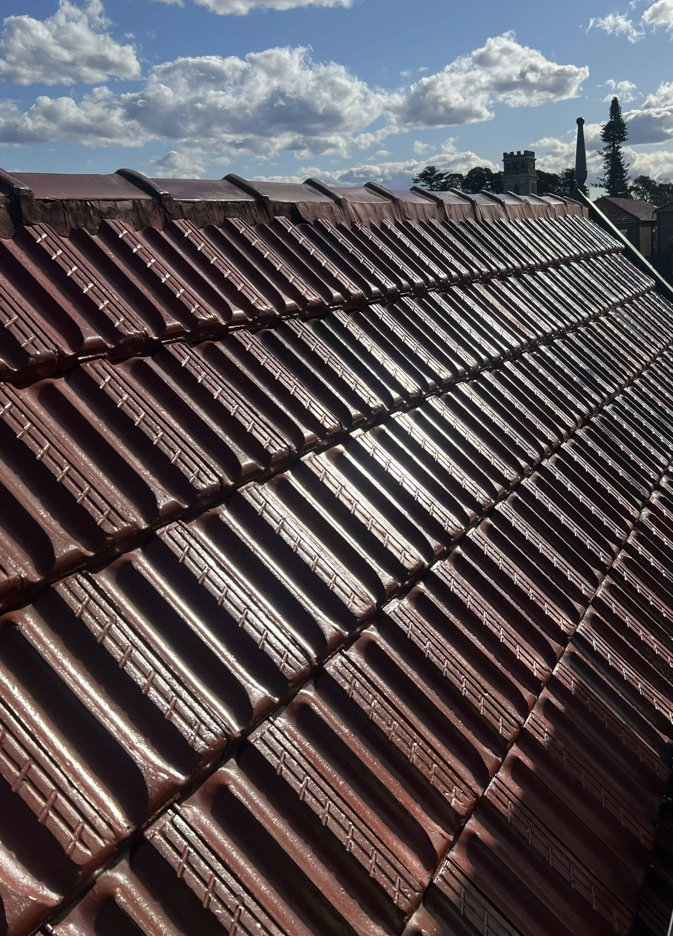 Close-up of a red clay tile roof with sunlight reflecting off the tiles, under a partly cloudy sky.