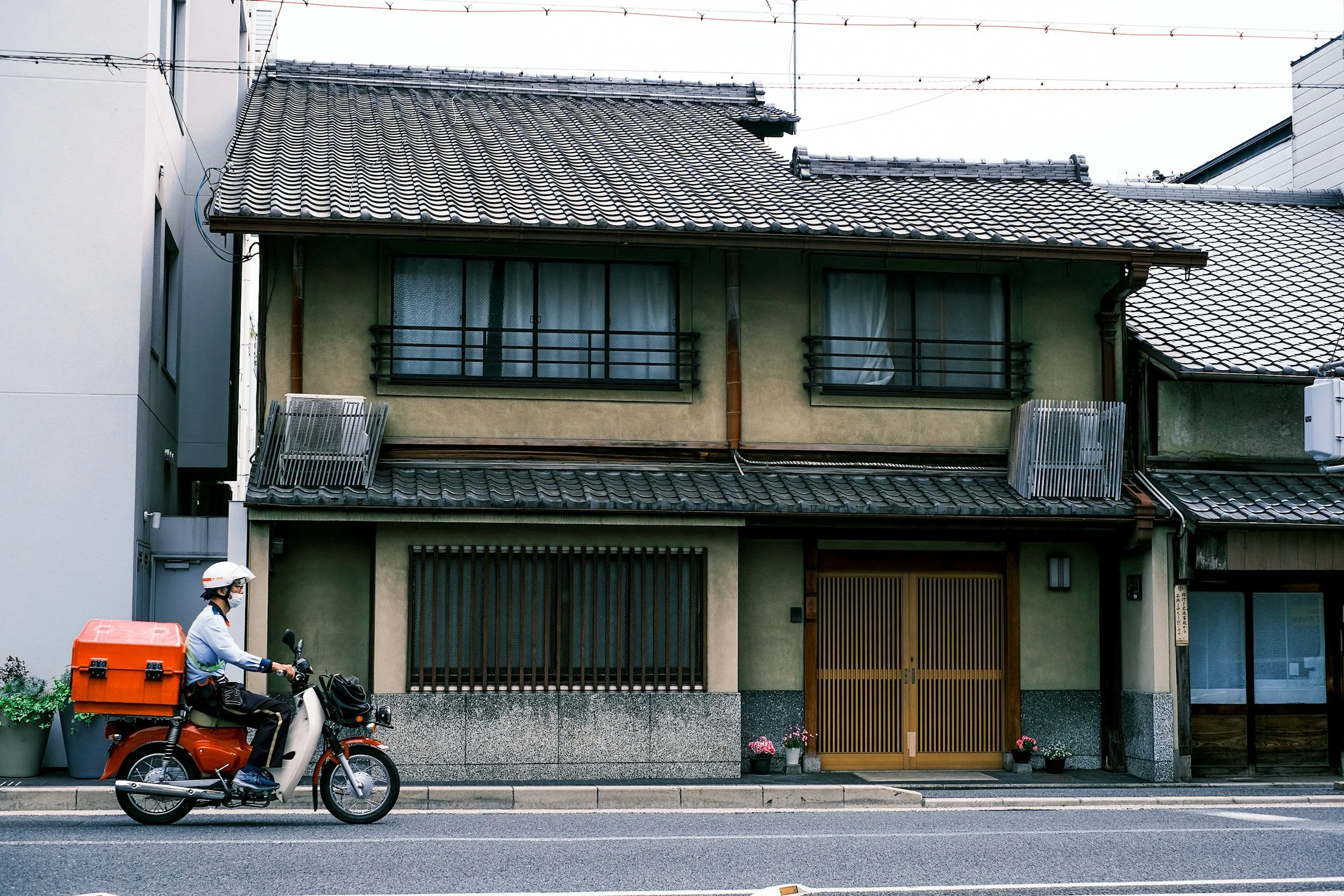 日本の古い住宅街にある瓦屋根の実家・空き家と、郵便配達員がバイクで通る日常風景。kokochiの調査対象となる築年数が経過した物件や管理のイメージ