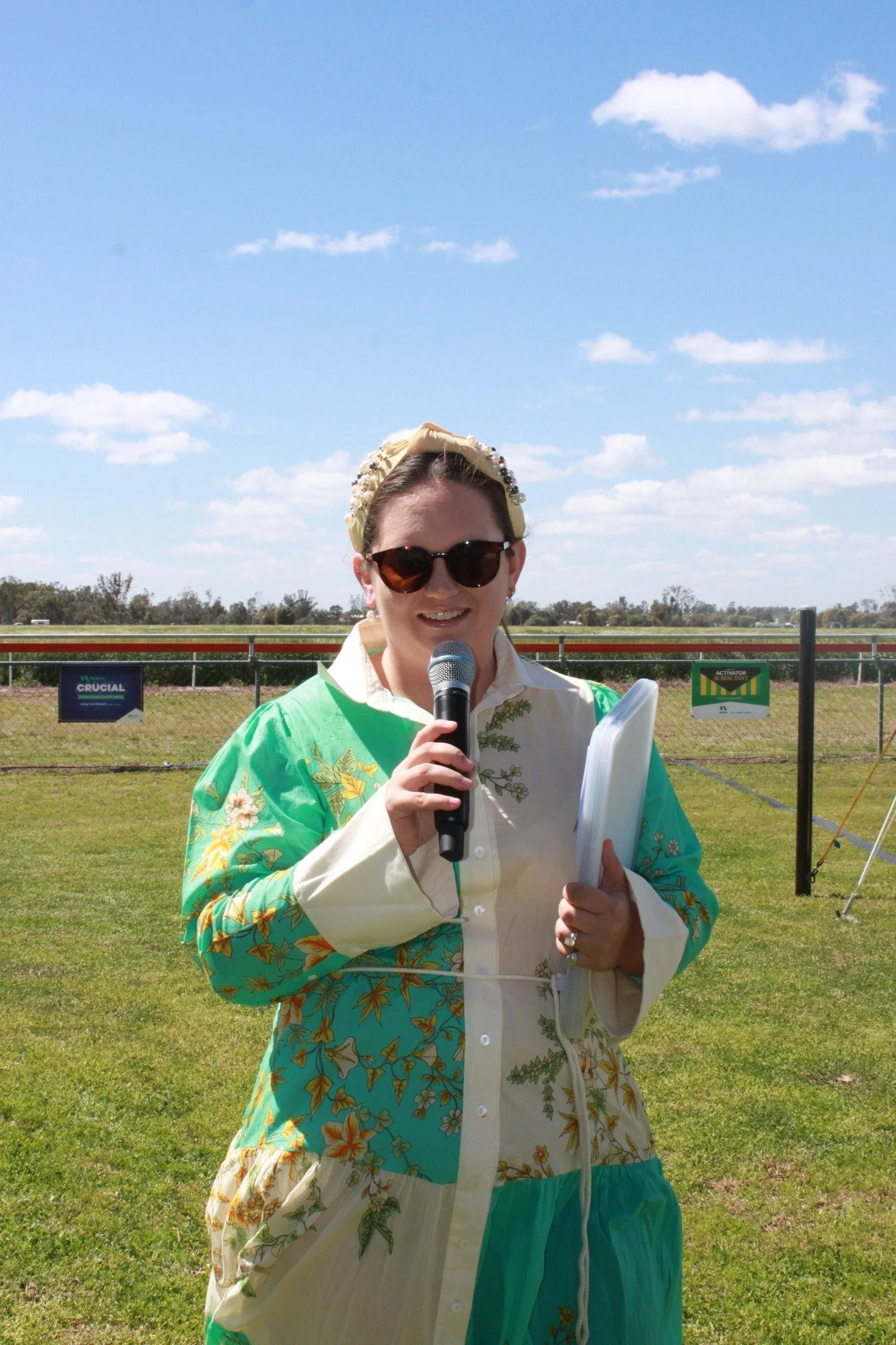 A woman wearing a floral dress, sunglasses, and a headband, standing outdoors on a grassy field under a partly cloudy sky, holding a microphone and some papers.