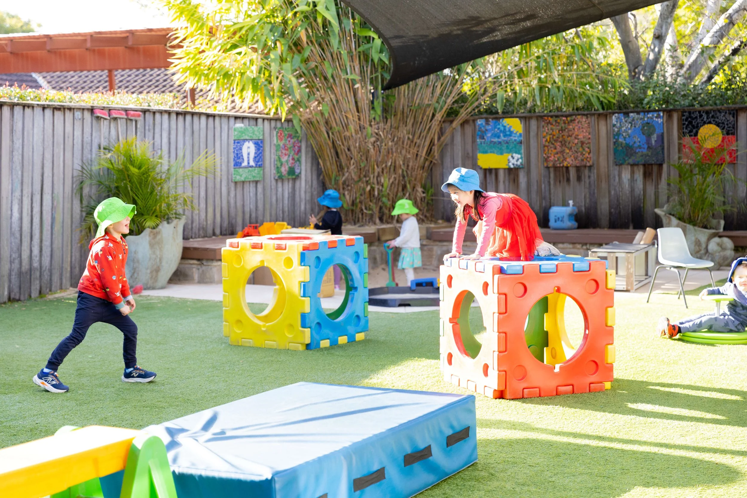 Children playing in a colorful outdoor playground with soft grass, large foam blocks, and toys, under a shaded area surrounded by a wooden fence and trees.