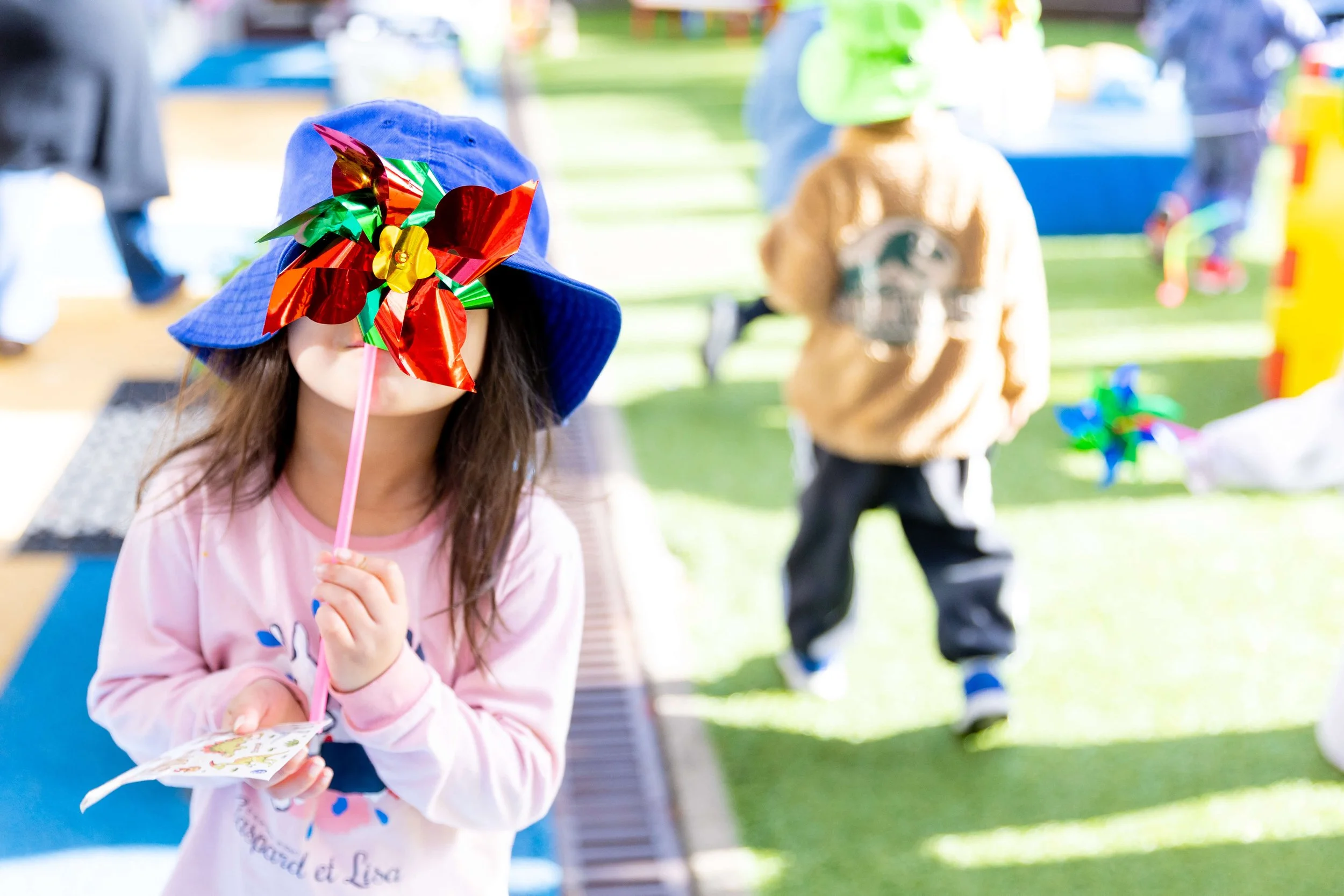 Child wearing a blue sun hat with a colorful pinwheel in front of her face, holding a card, at an outdoor event with children in the background.