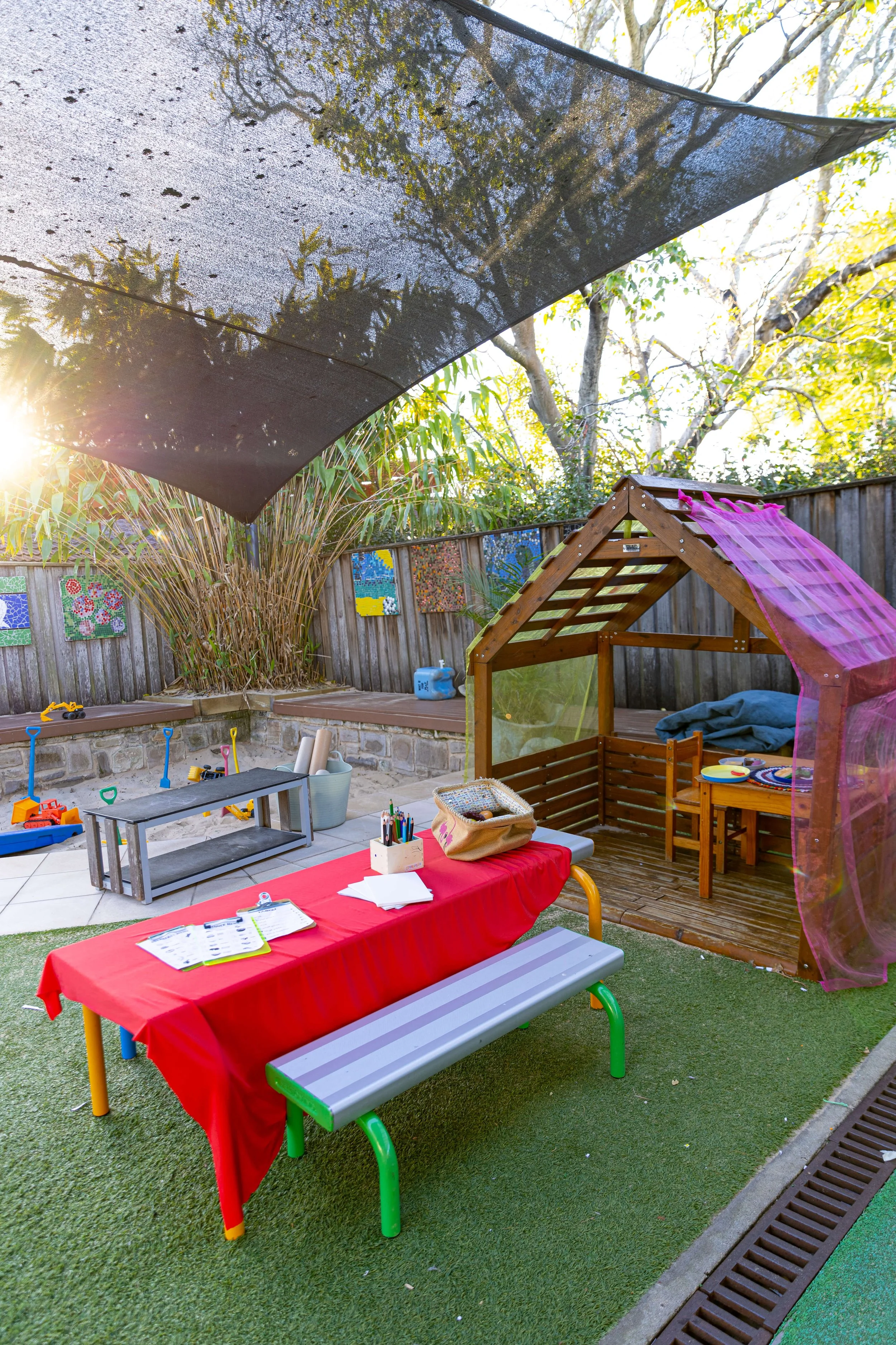 Children's outdoor play area with a red-clothed table, art supplies, a wooden sandbox with pink curtains, and a shaded yard with toys and artwork on a wooden fence.