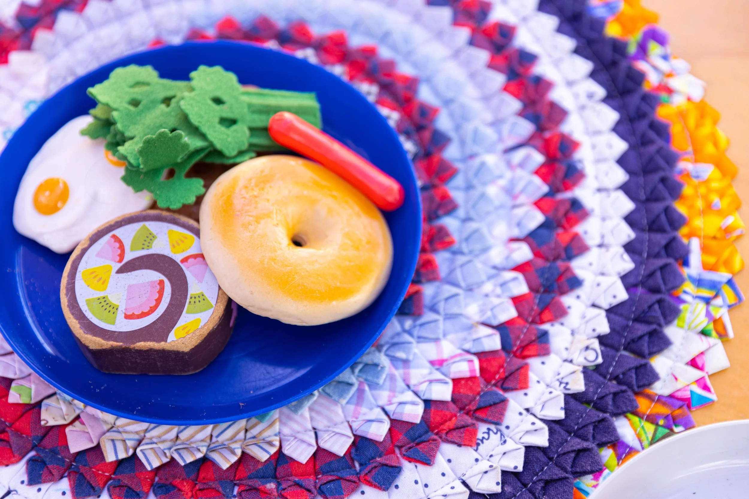 Colorful plate with pretend food including a fried egg, a bagel, felt vegetables, and a sausage, placed on a woven, multicolored placemat.