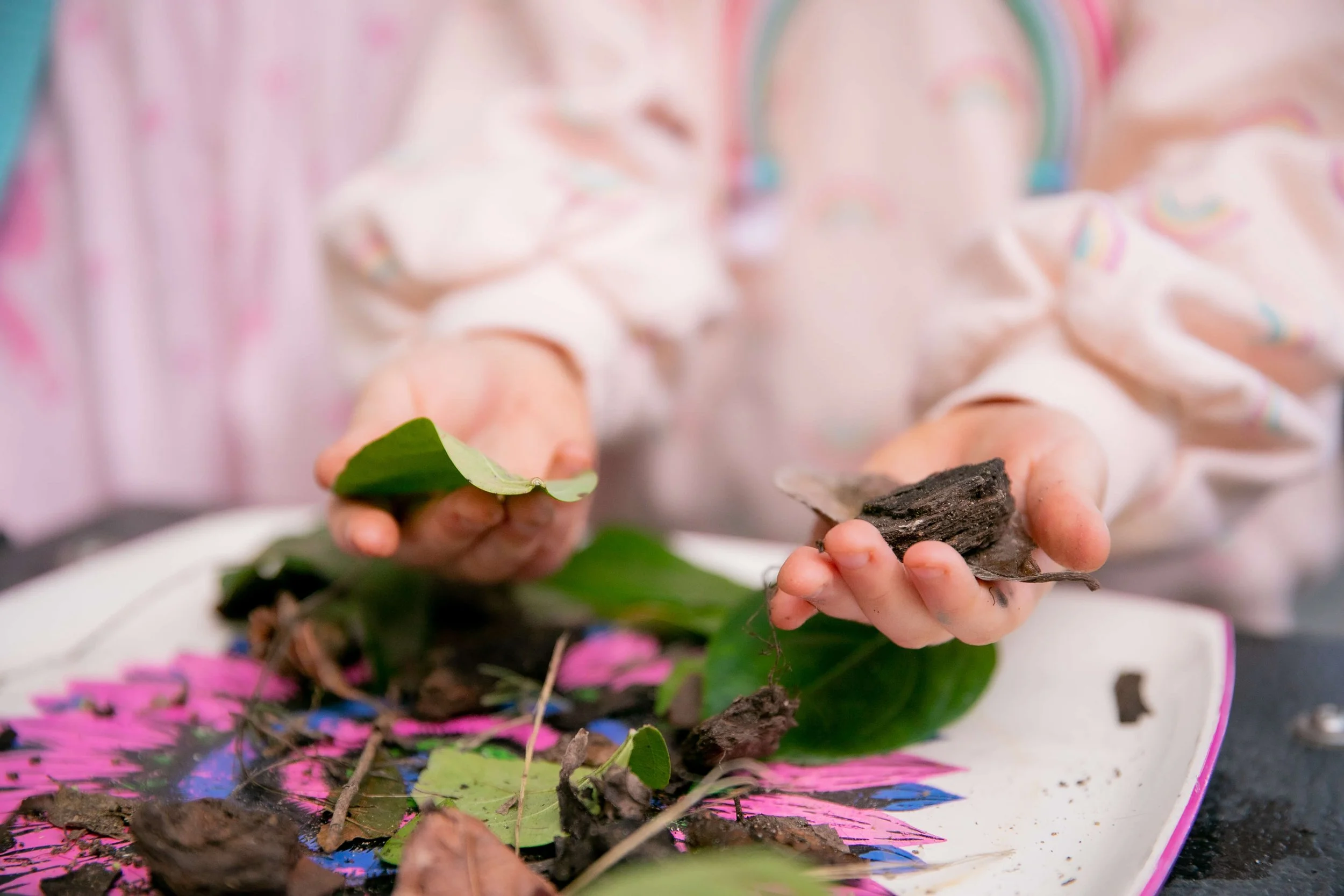 Child holding soil and plant debris over a decorative plate with leaves, sticks, and dirt.