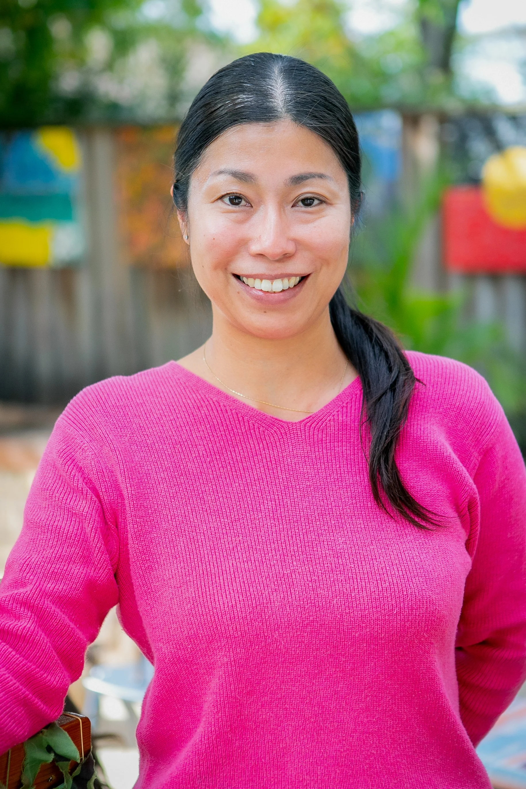 Portrait of a smiling woman with long dark hair, wearing a pink sweater, standing outdoors with colorful blurred background.