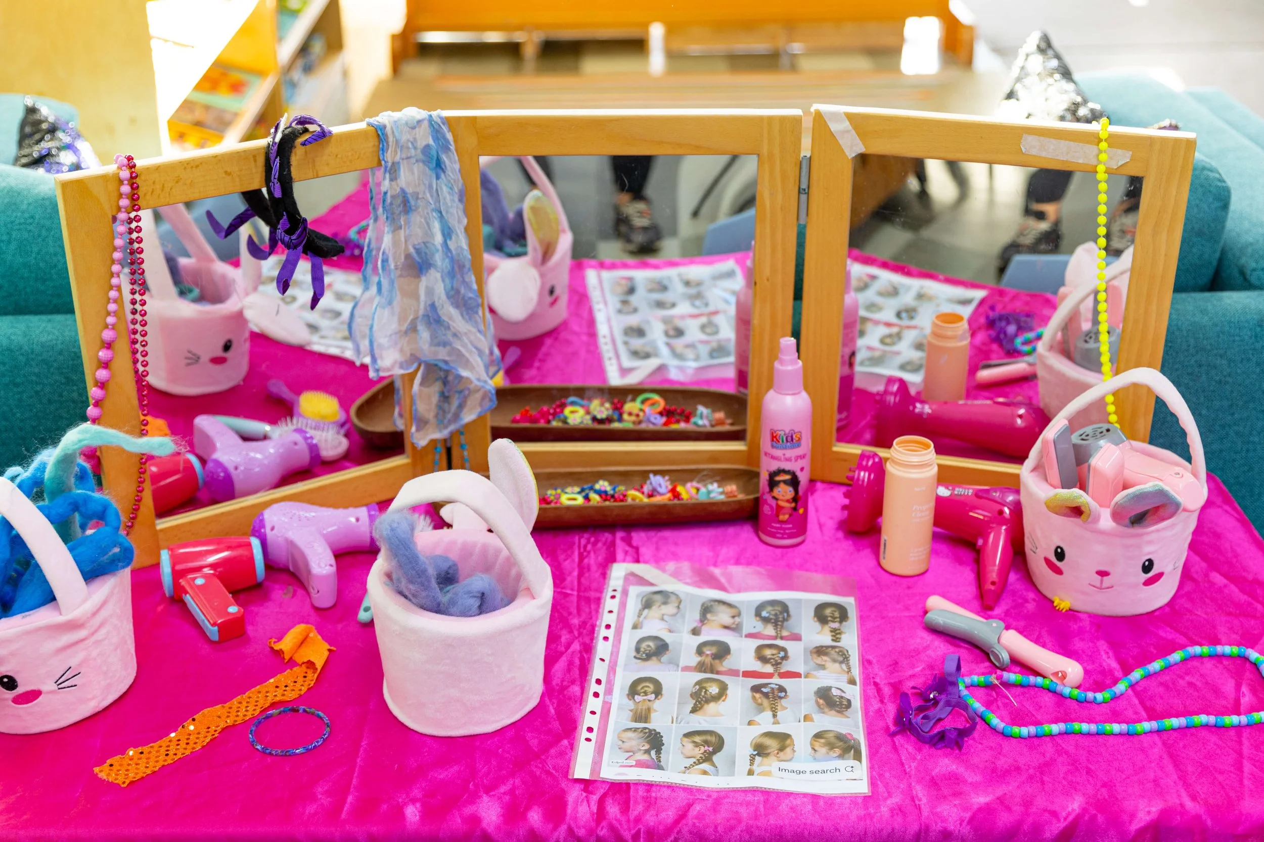 A table with various hair styling tools, accessories, and hair clips, set up for a children's hair braiding activity. The table has pink fabric and mirrors, with baskets filled with colorful hair ties and scarves, along with spray bottles and toy hai
