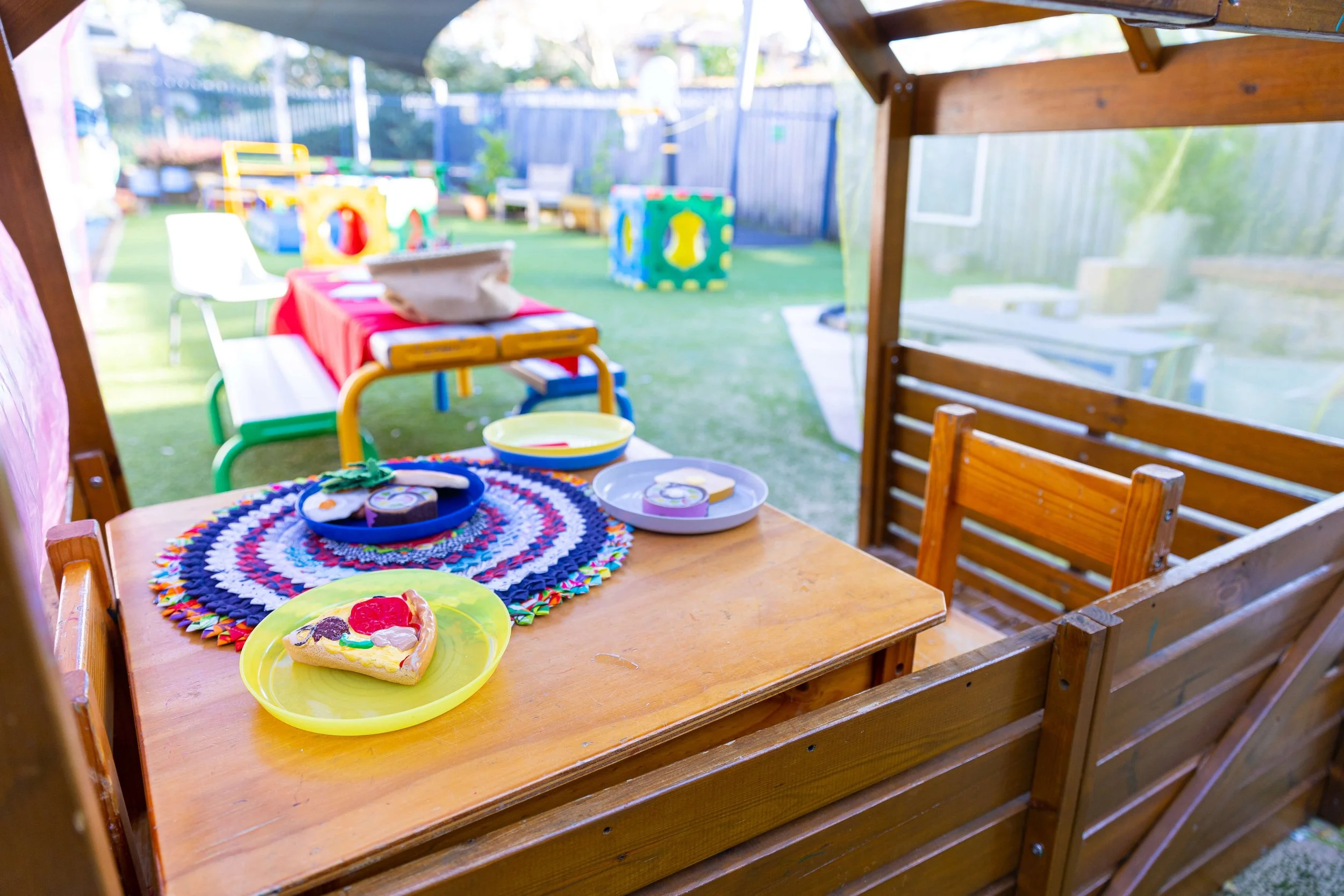 Outdoor playground with colorful play structures, tables, and chairs, with a wooden table in the foreground holding a decorated cake and party supplies.