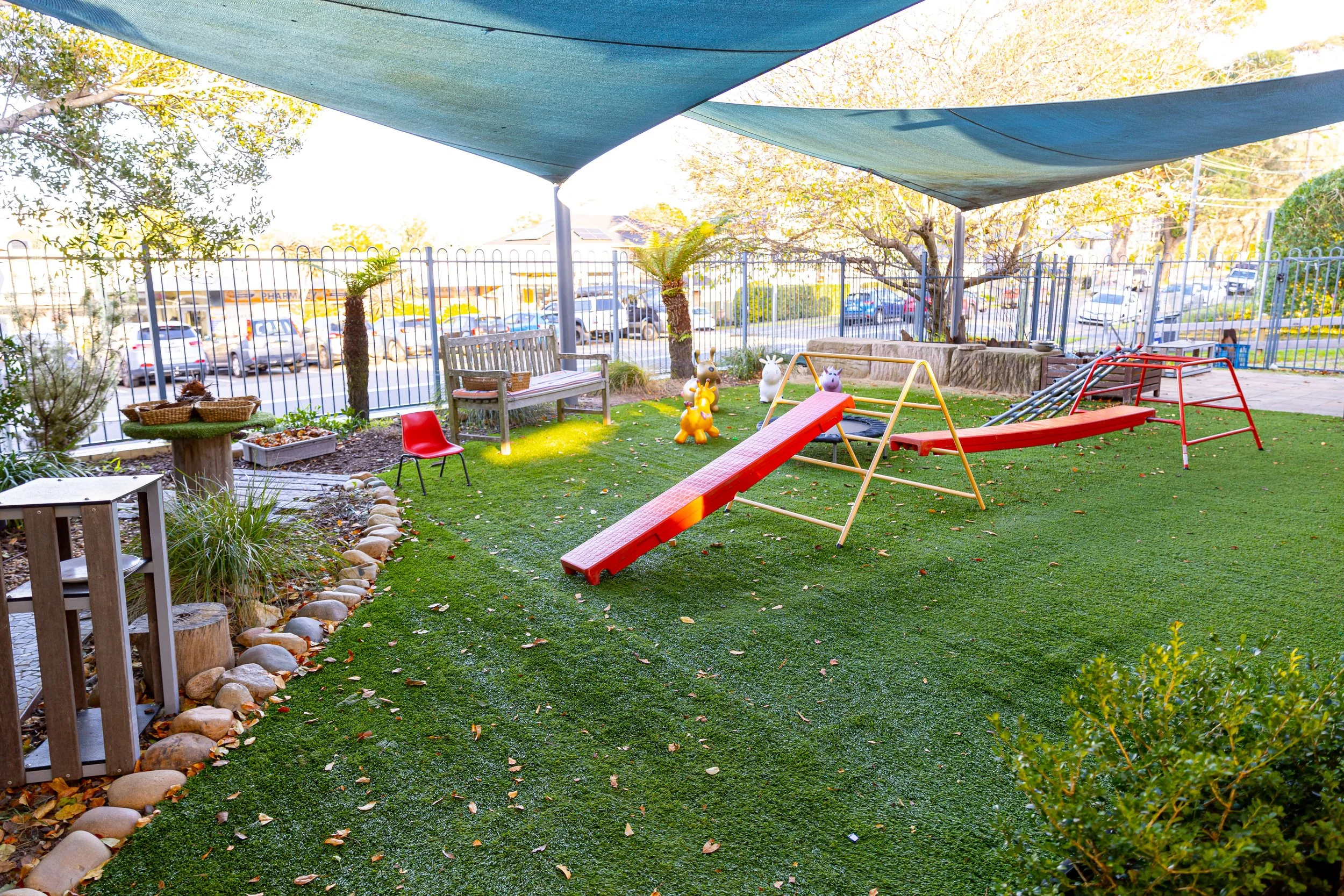 Children's outdoor playground with a slide, climbing structure, and animal-shaped playground equipment, shaded by a blue canopy, surrounded by trees, bushes, and a fence.
