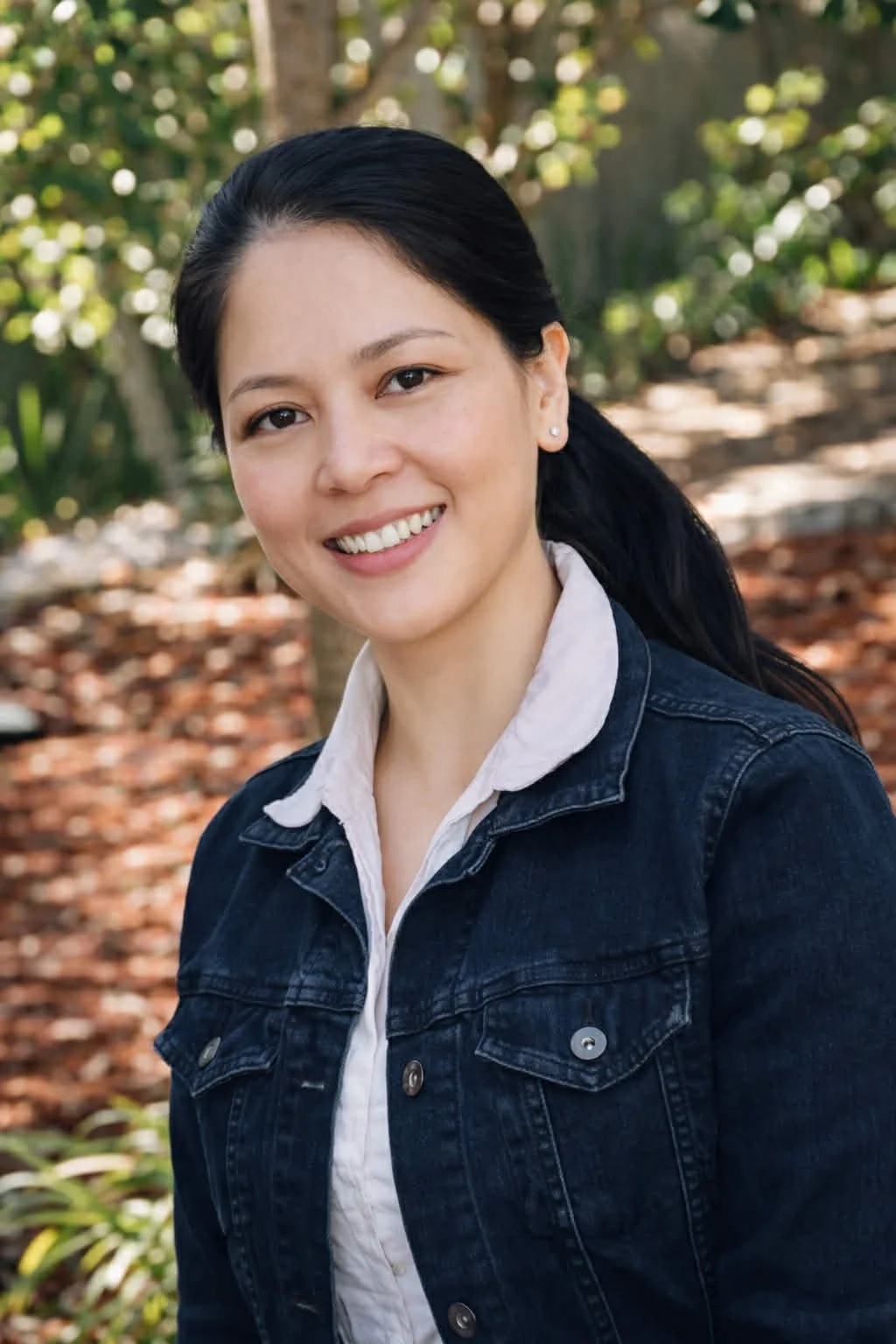 Young woman with dark hair in a ponytail smiling outdoors, wearing a white shirt and denim jacket, with a background of trees and fallen leaves.