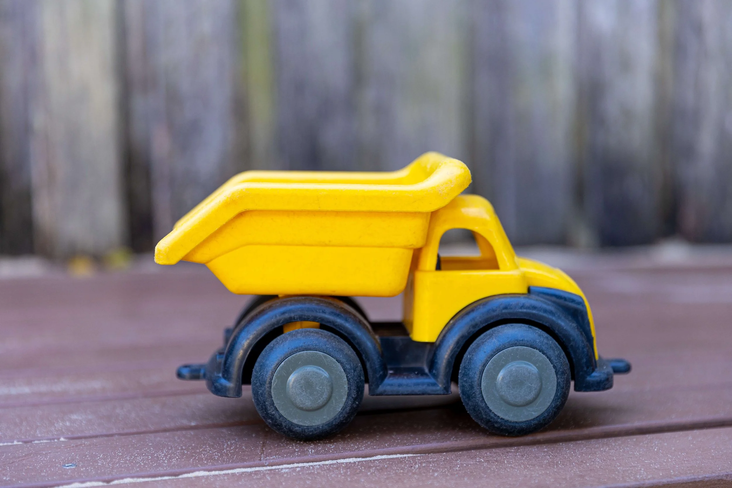 A yellow toy dump truck with a yellow bed, black wheels, and a yellow cab, placed on a wooden surface with a blurred outdoor background.