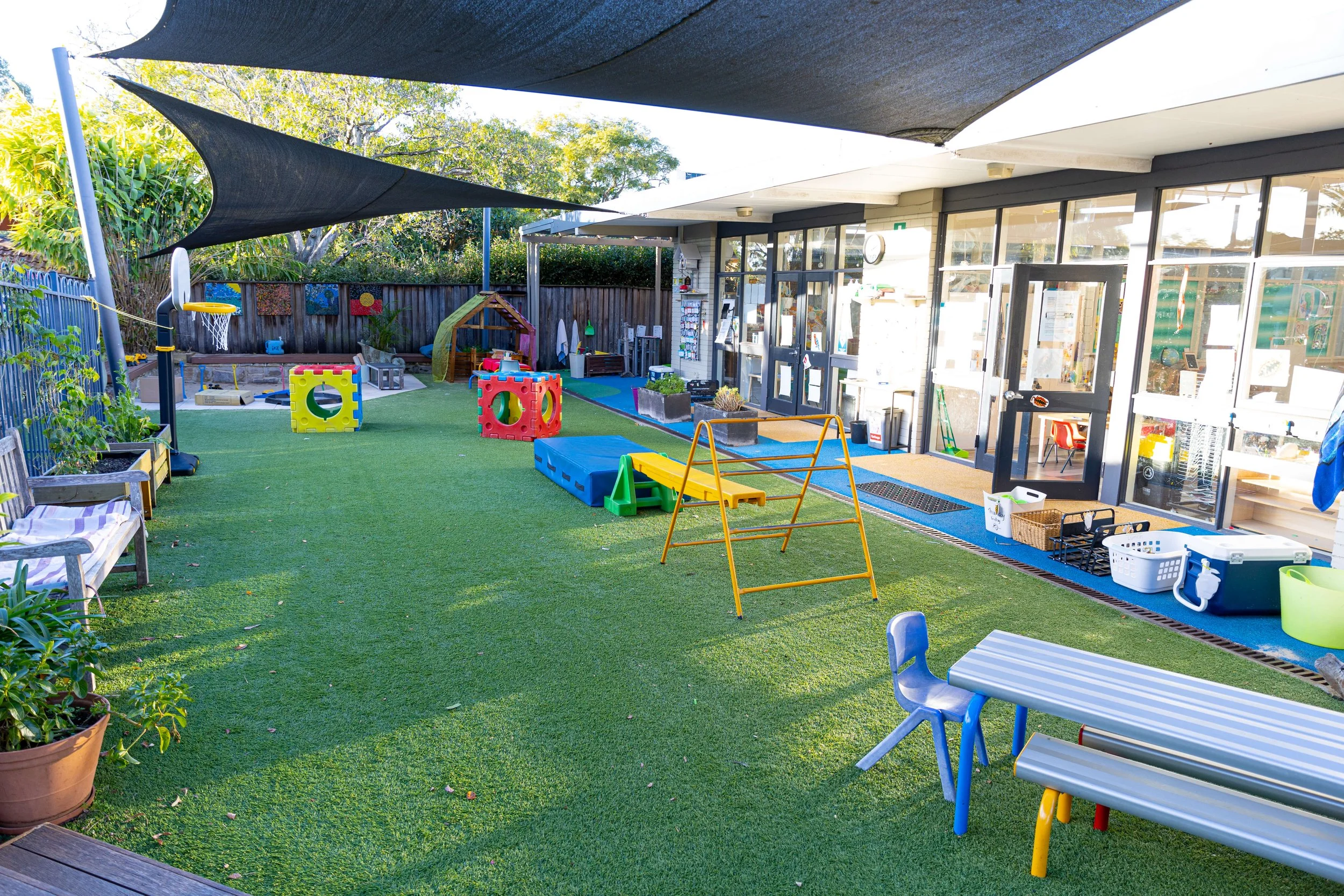 Outdoor preschool playground area with artificial grass, colorful play equipment, shade sails, and a large windowed building in the background.