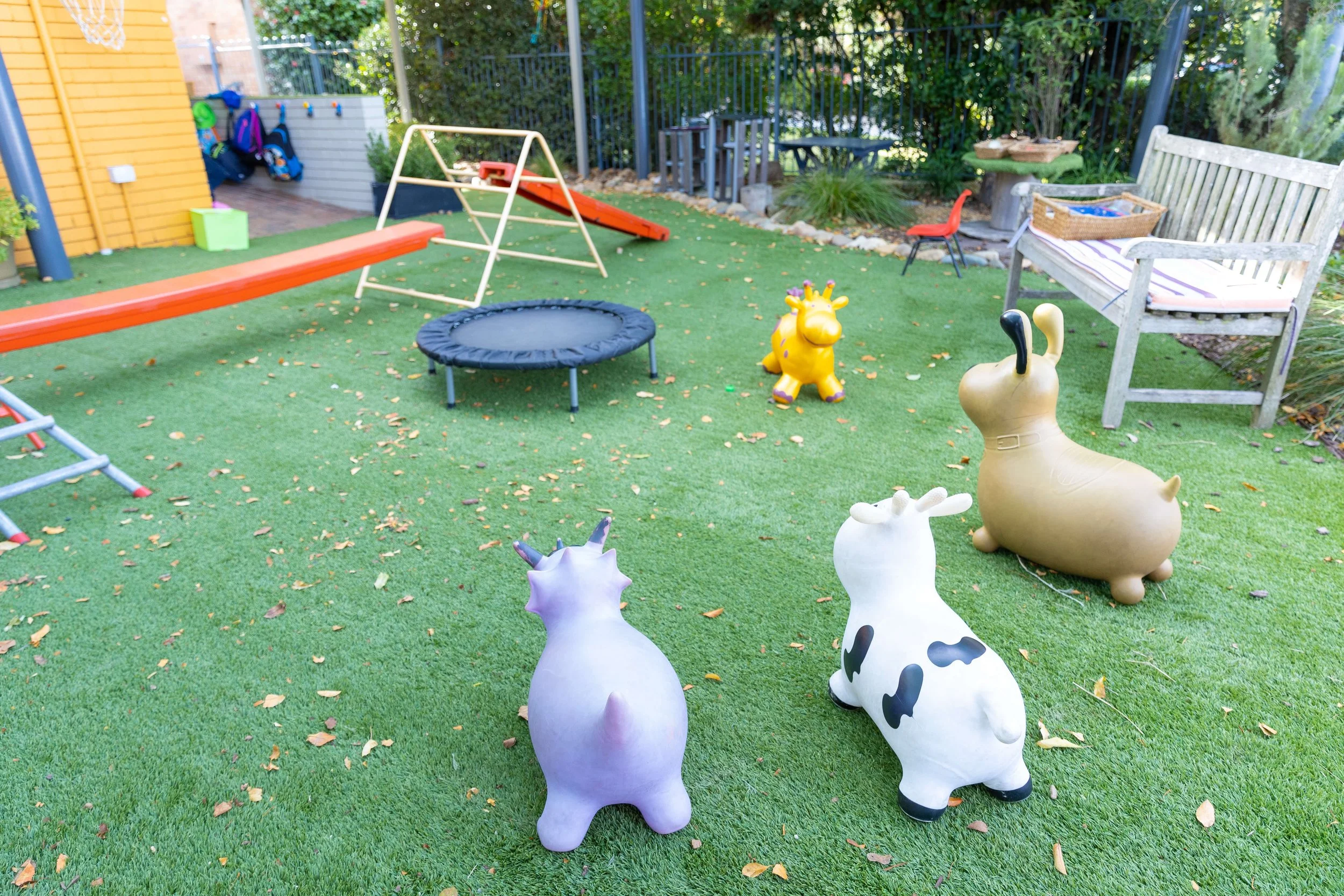 Children's outdoor play area with various animal-shaped and cartoon-like garden ornaments, a trampoline, a slide, a bench, and toys on artificial grass.