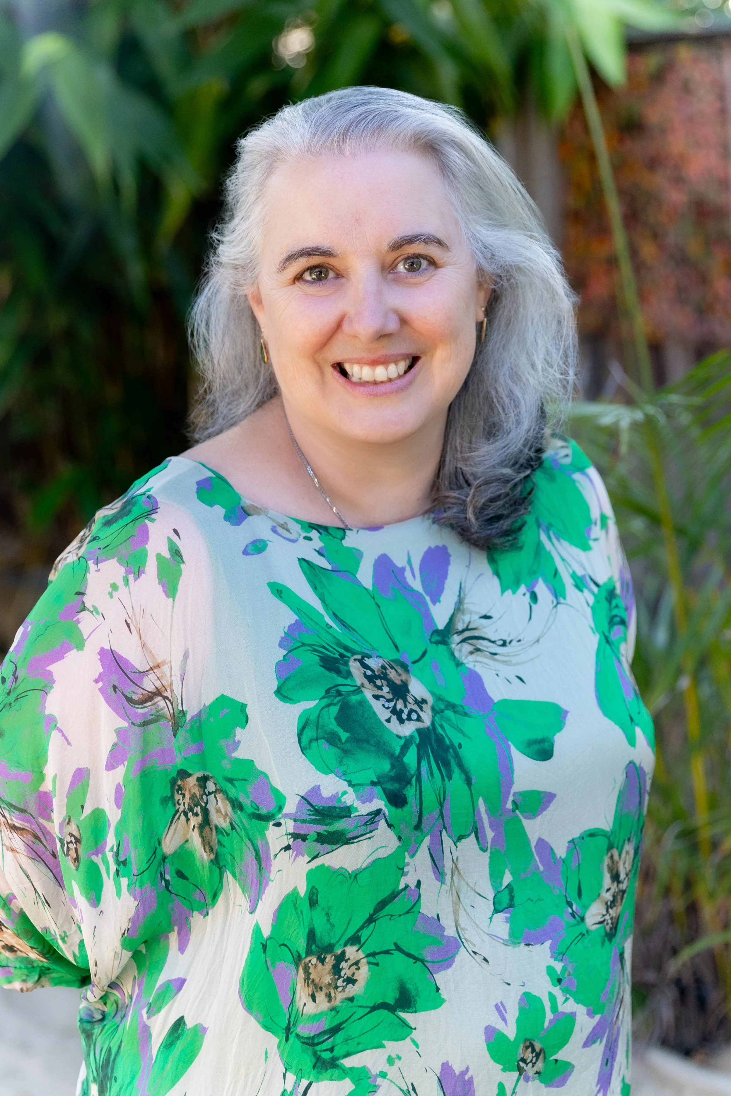Woman with gray hair and smiling outdoors, wearing a blue top and a light gray scarf, with a wooden fence and trees in the background.