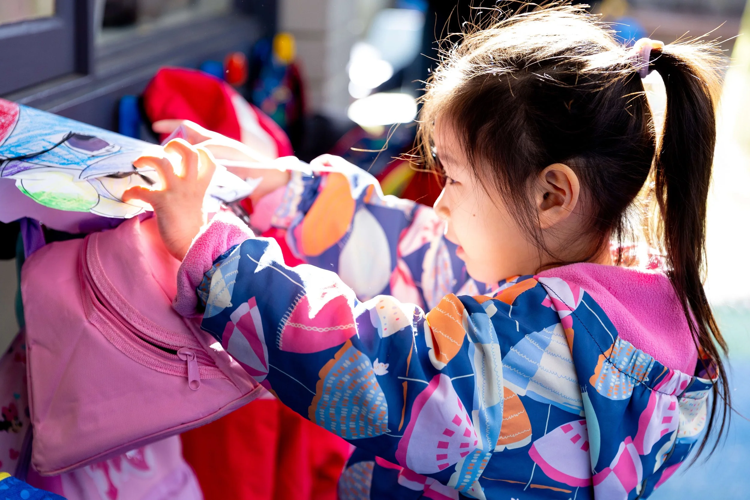 Young girl with a ponytail and colorful jacket hanging artwork on a pink backpack.