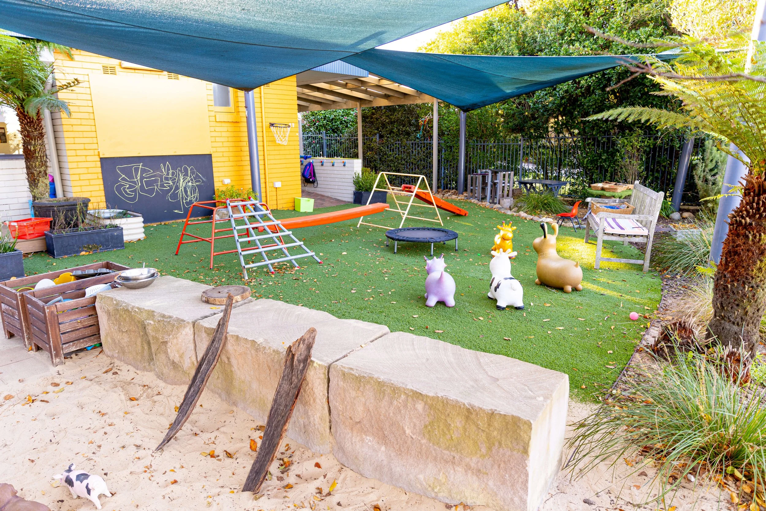 Colorful outdoor play area with artificial grass, a shaded canopy, and various playground equipment including a small trampoline, slide, and climbing frame. There are animal-shaped ride-on toys, benches, plants, and graffiti on the yellow building wa