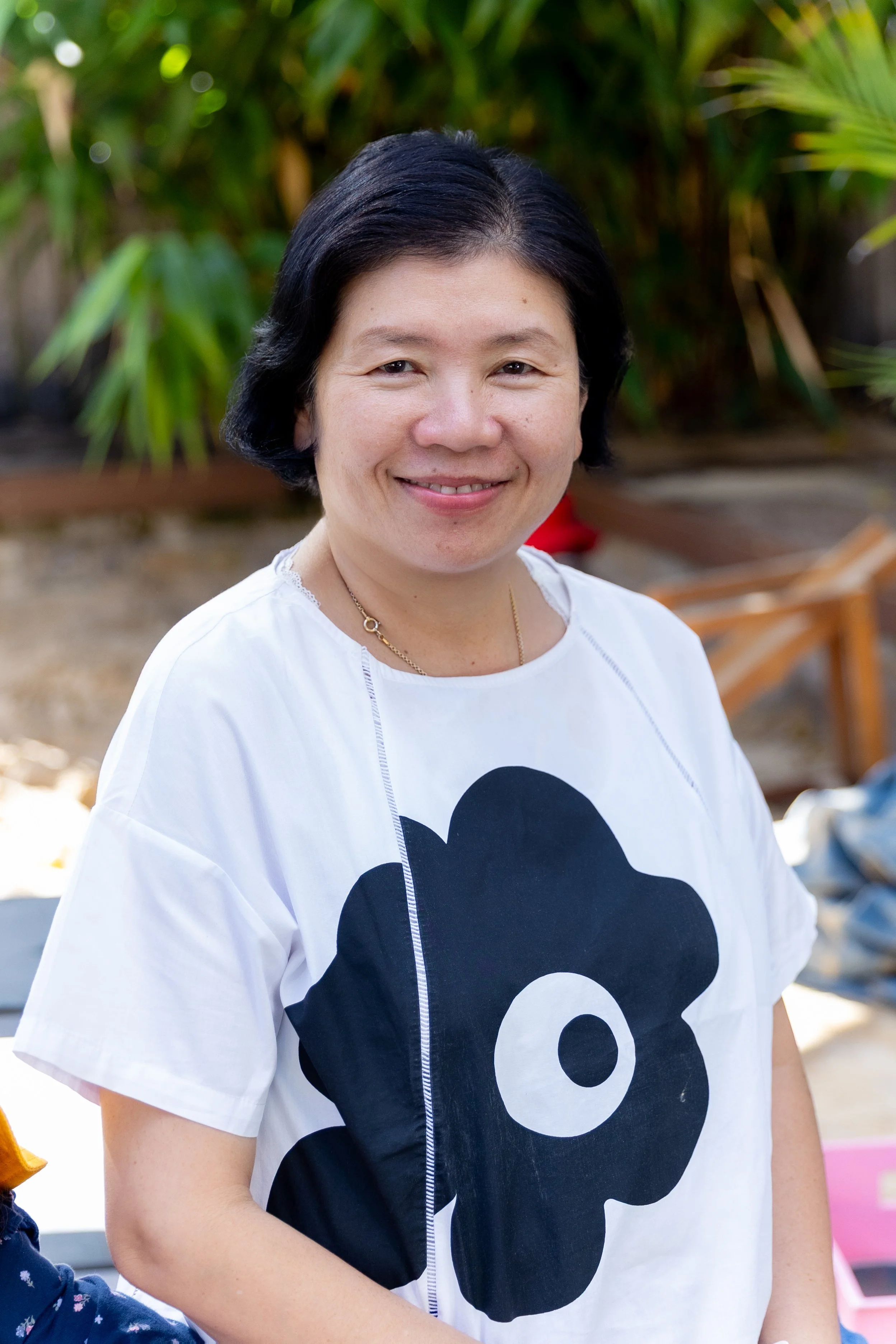 A woman smiling outdoors, wearing a red shirt with a small white design on the pocket. She has short black hair, a watch on her left wrist, and is holding some green leafy foliage. Trees and a wooden fence are in the background.