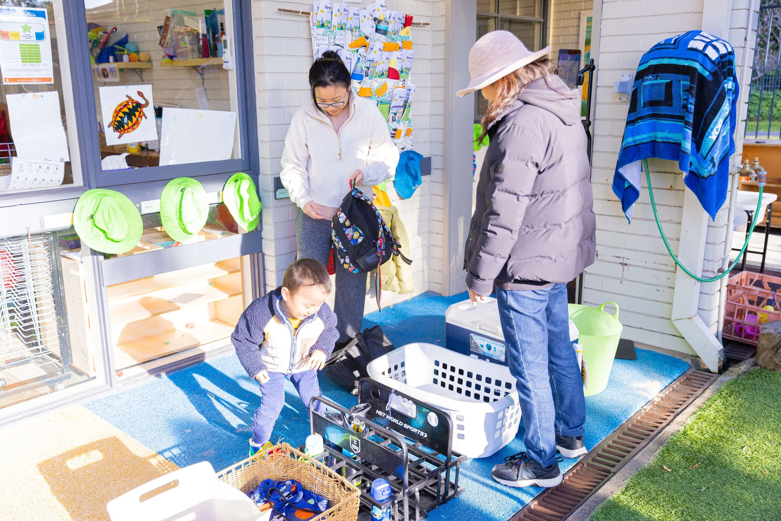 Two women and a young boy packing or unpacking items outside a building. The women are wearing jackets, one with a hood and hat, and the boy is looking down at something. There are baskets and containers filled with belongings, possibly for a trip or