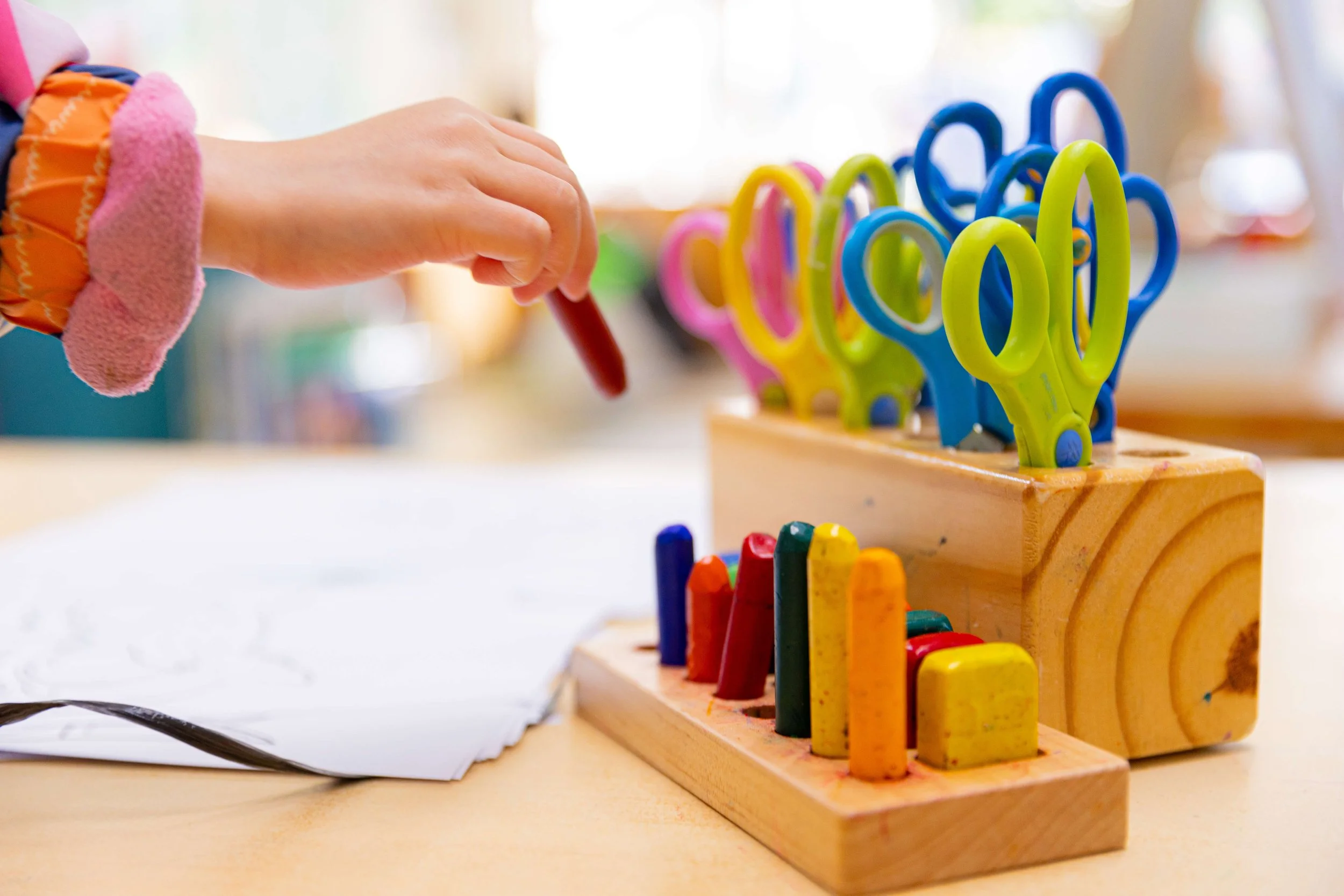Child's hand reaching for a red crayon from a wooden box filled with colorful crayons, with another wooden box containing brightly colored scissors nearby.