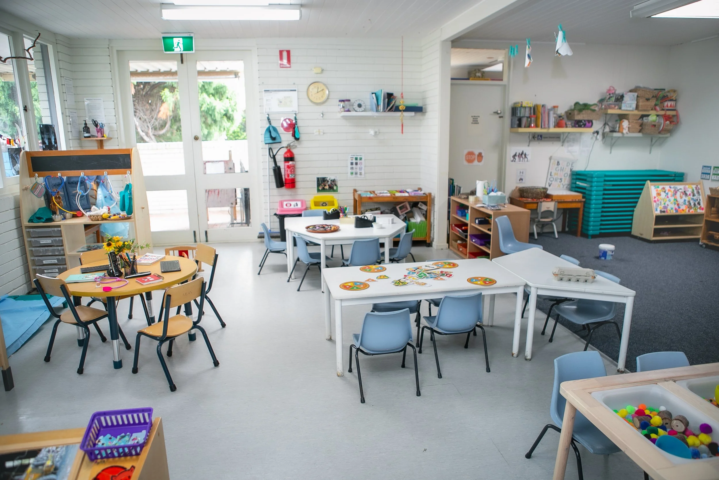 An empty preschool classroom with small tables, chairs, and various toys and educational materials, with sunlight coming through glass doors.
