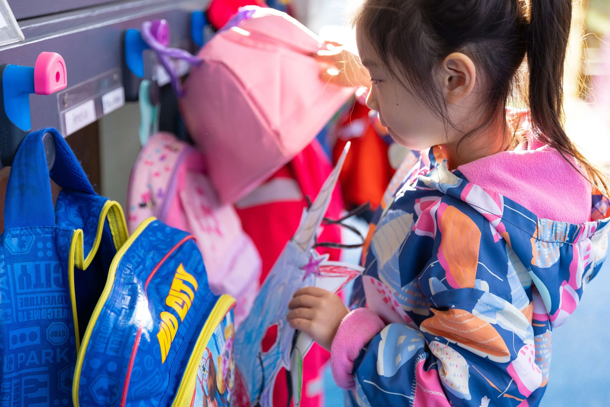 Young girl in colorful jacket looking at a pink and blue backpack with a note in a school hallway.