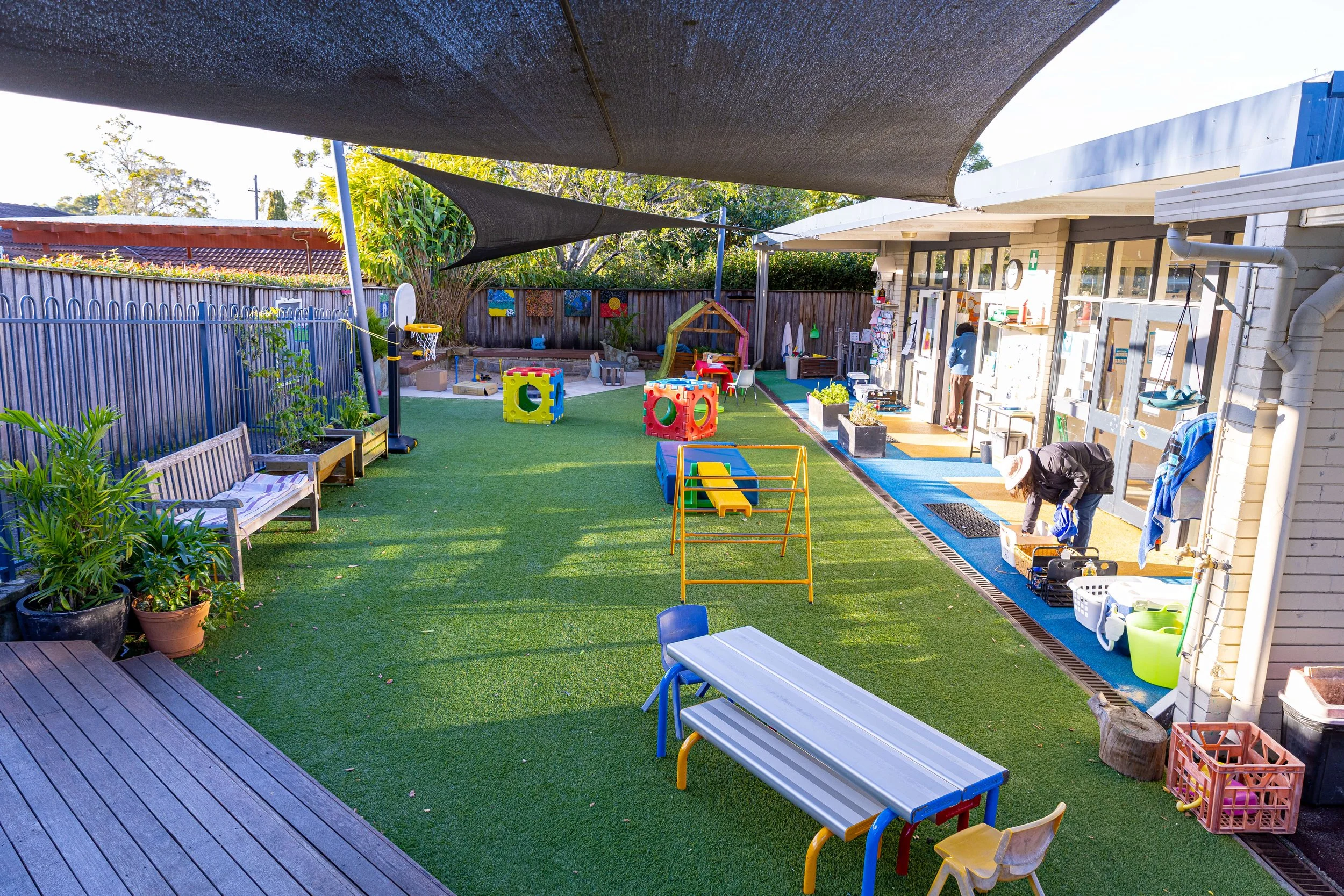 Outdoor daycare playground with artificial grass, benches, potted plants, colorful toys, a small slide, and two adults organizing items next to a childcare building under shade sails.
