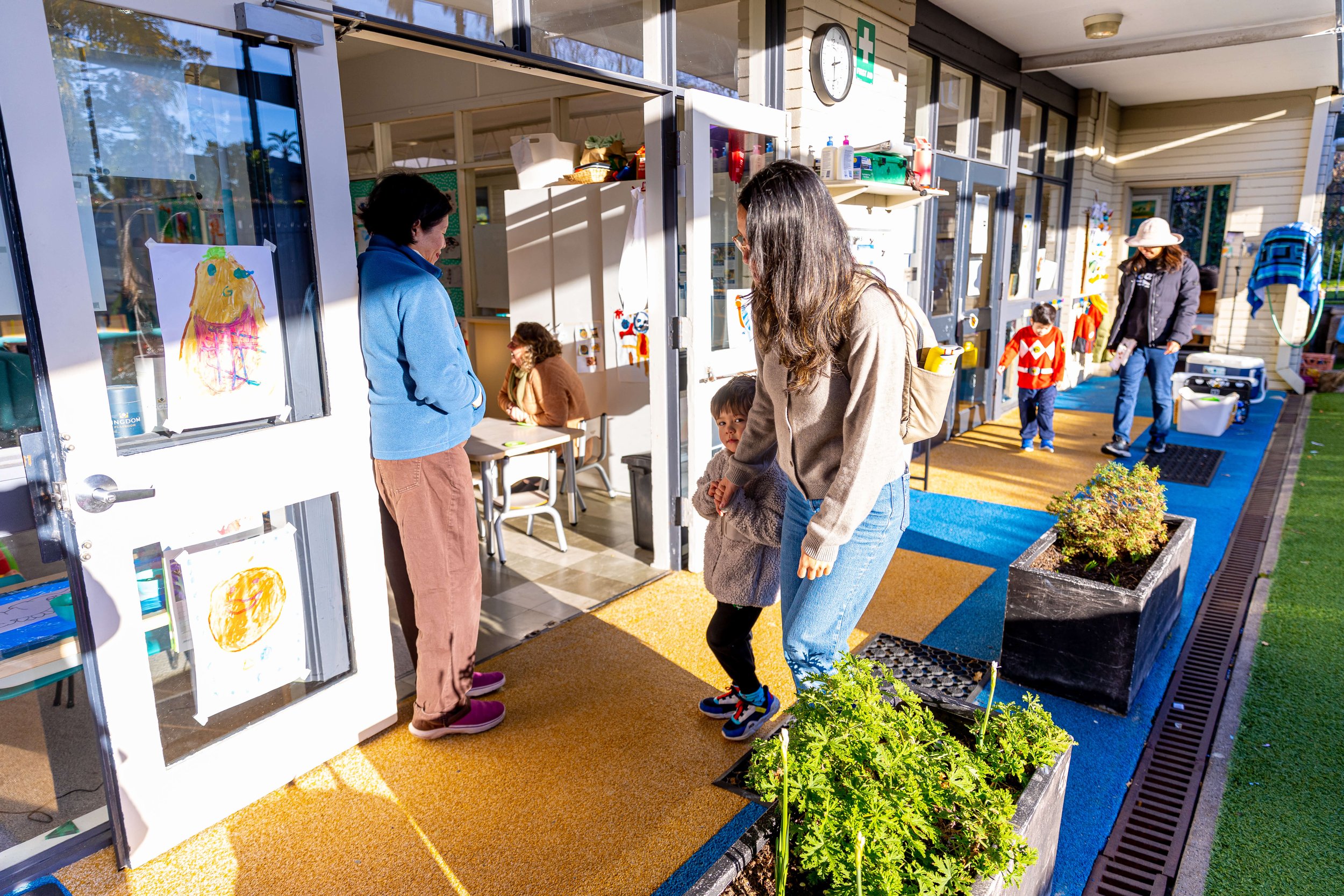 People entering a colorful school or community center with children and adults outside, sunlight shining on the scene, and artwork displayed on the doors.