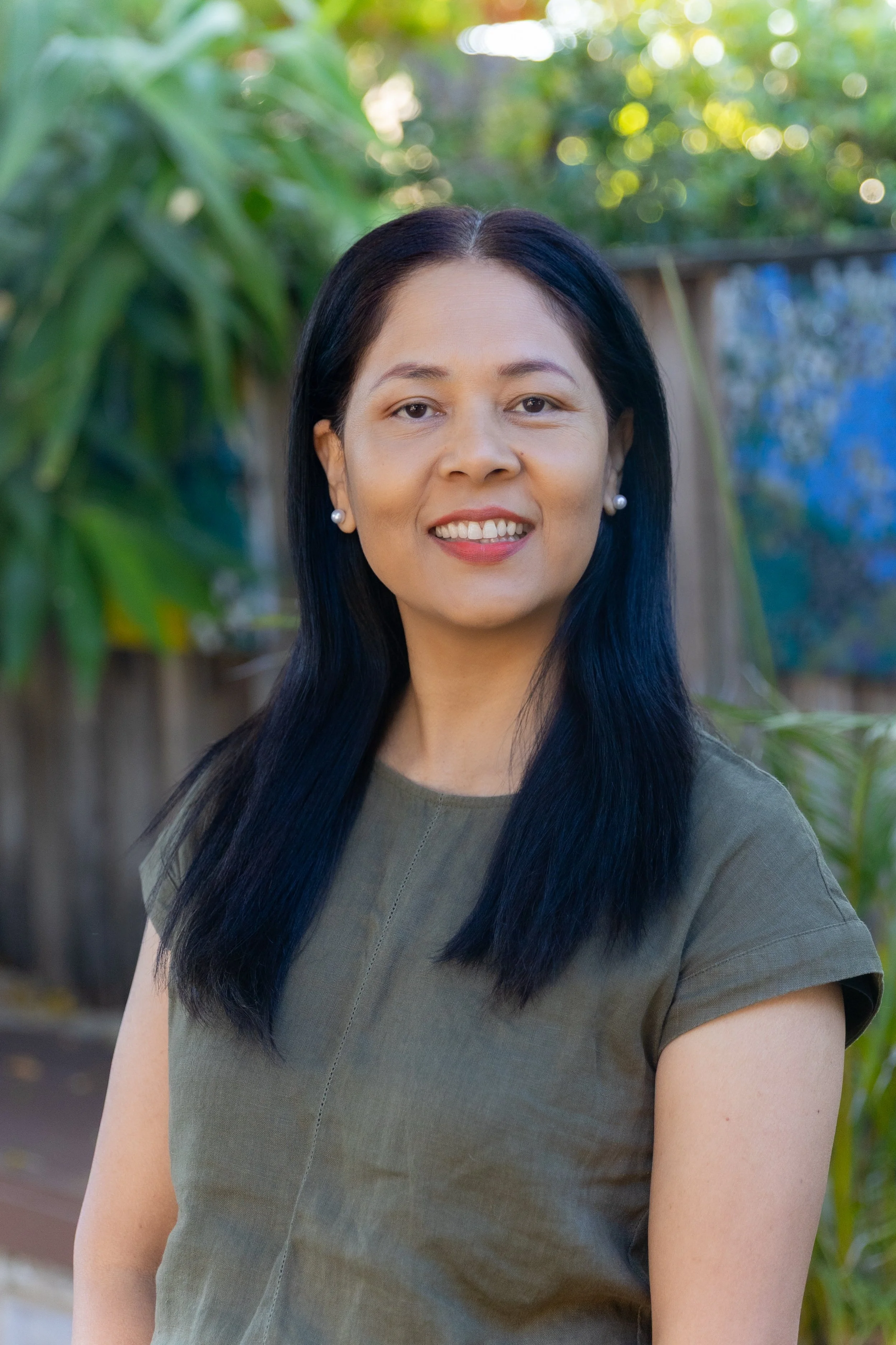 Young woman with dark hair in a ponytail smiling outdoors, wearing a white shirt and denim jacket, with a background of trees and fallen leaves.
