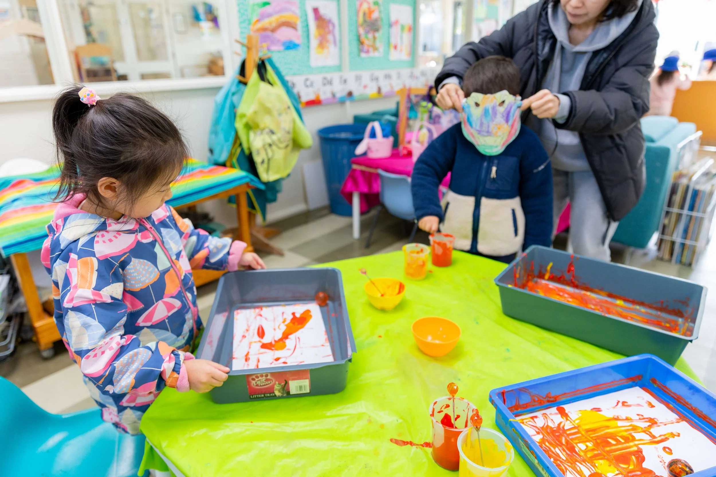 Young girl and boy participating in finger painting activity at a colorful classroom table with paint containers and paper, supervised by an adult.