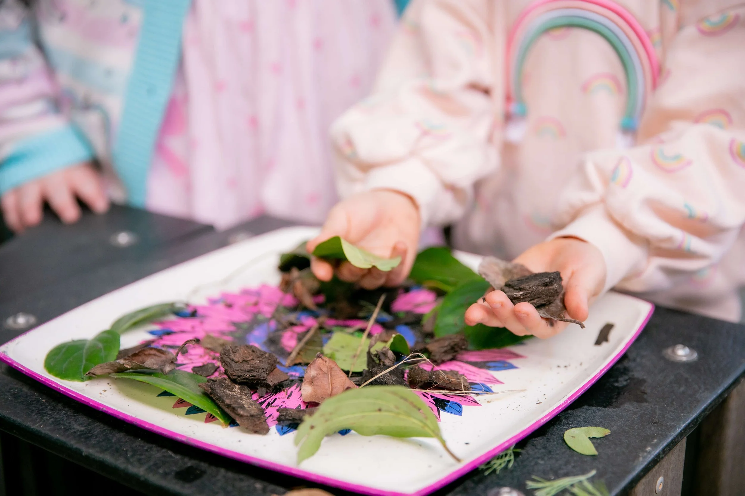 Child's hands showing soil, leaves, and small pieces of bark or wood on a colorful tray with a pink and black floral pattern.