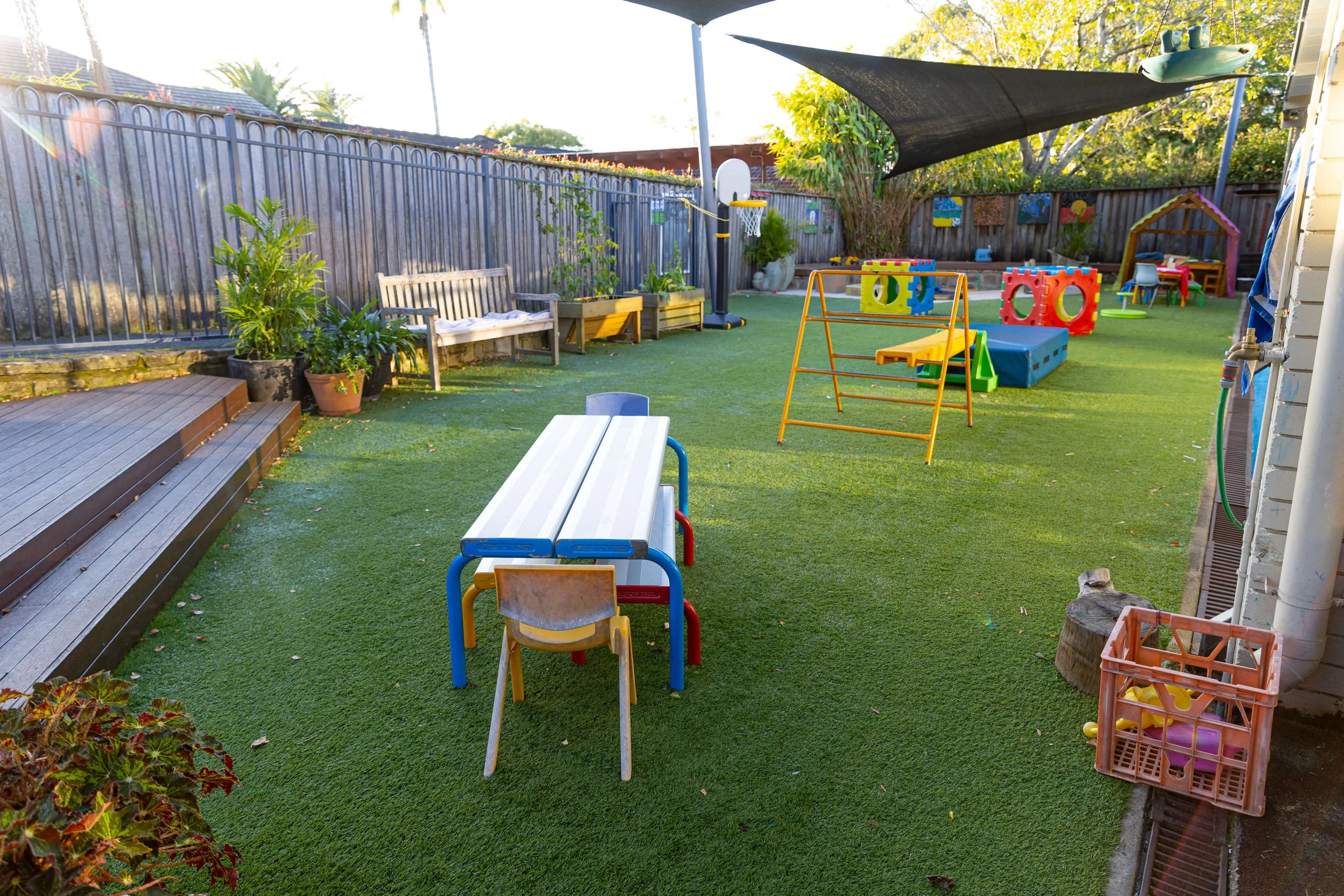 Children's outdoor playground with artificial grass, play structures, toys, benches, plants, and shade sail.