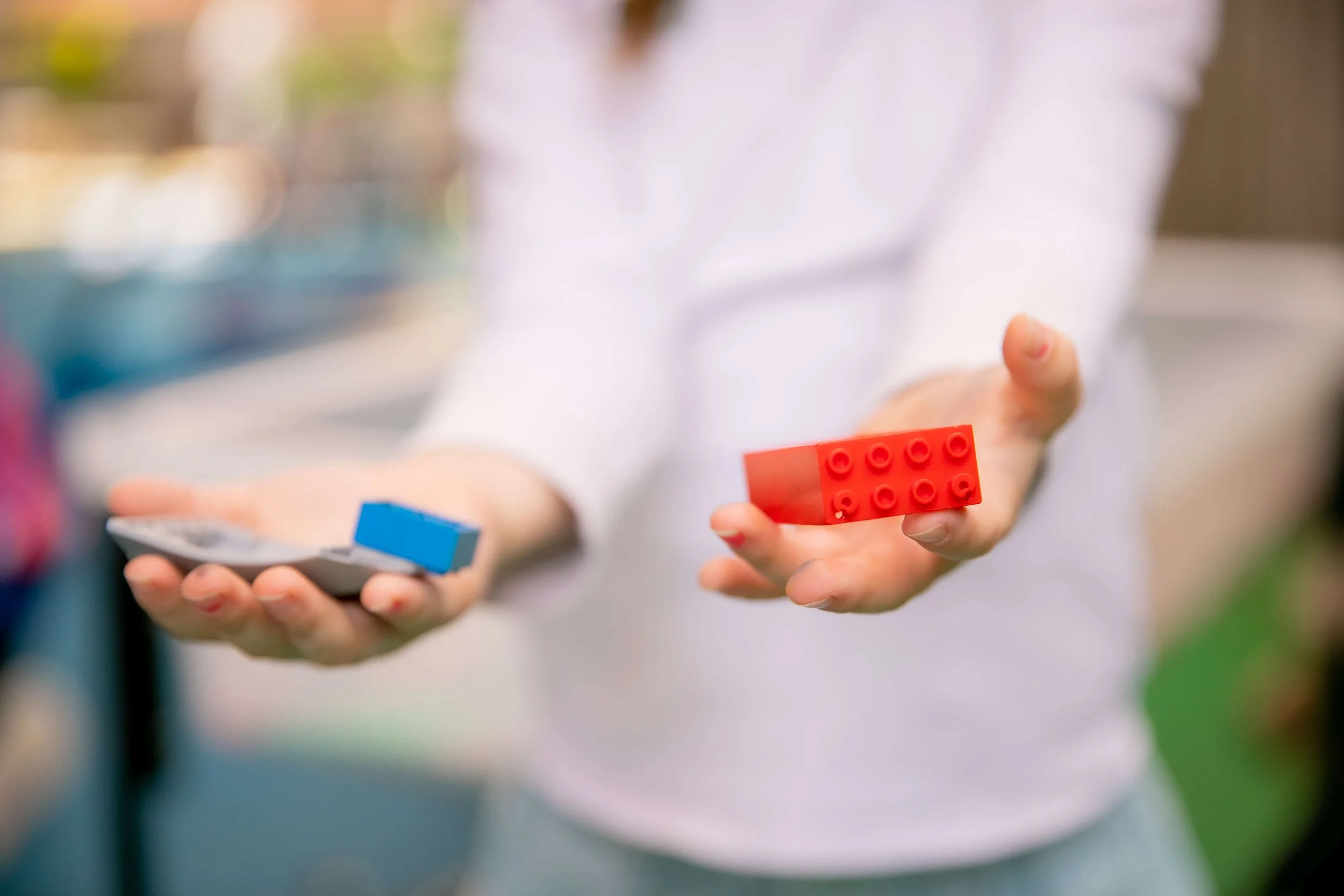 Person holding a red LEGO brick in one hand and a gray device with a blue piece in the other hand, outdoors.