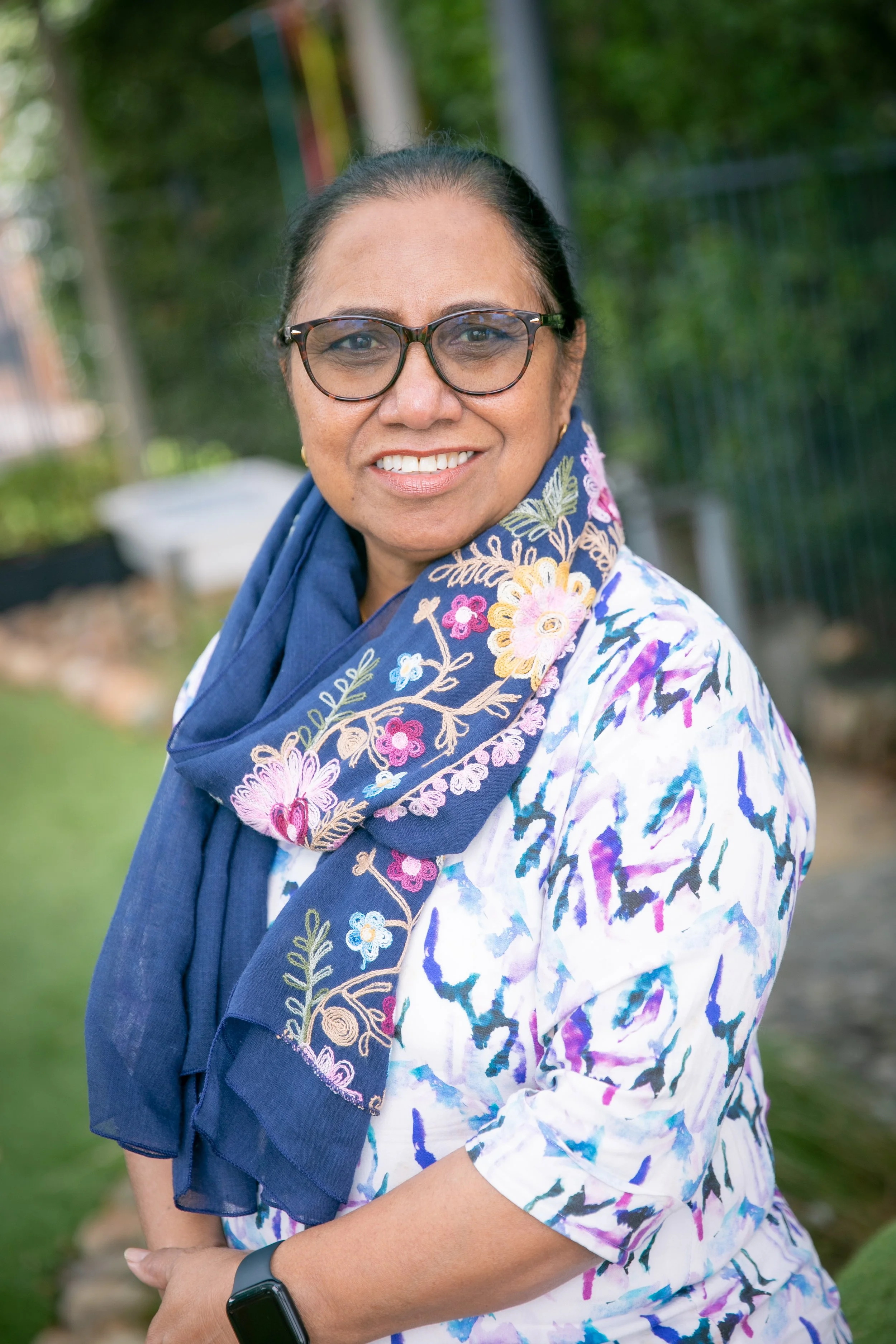 A woman with dark hair, glasses, and a colorful scarf smiling outdoors in a garden or park setting.