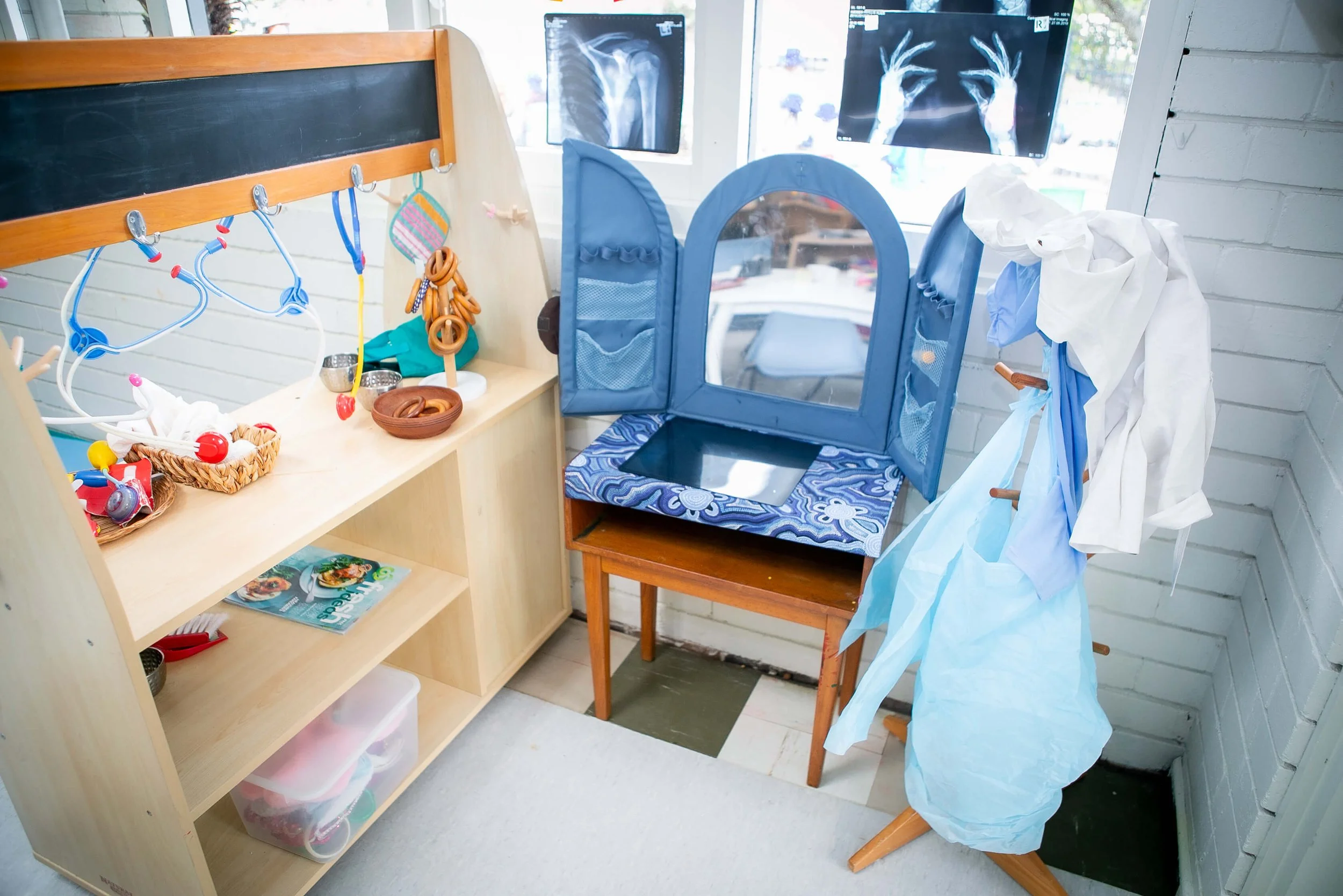 A play area with toy kitchen accessories, a small mirror, and costumes, set against a window with X-ray images displayed on monitors.