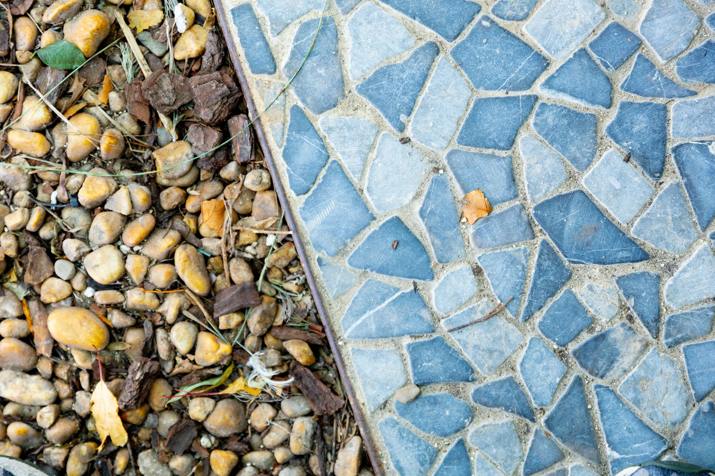 Close-up image of a transition between small yellow pebbles and a mosaic of irregular blue tiles separated by gray grout.