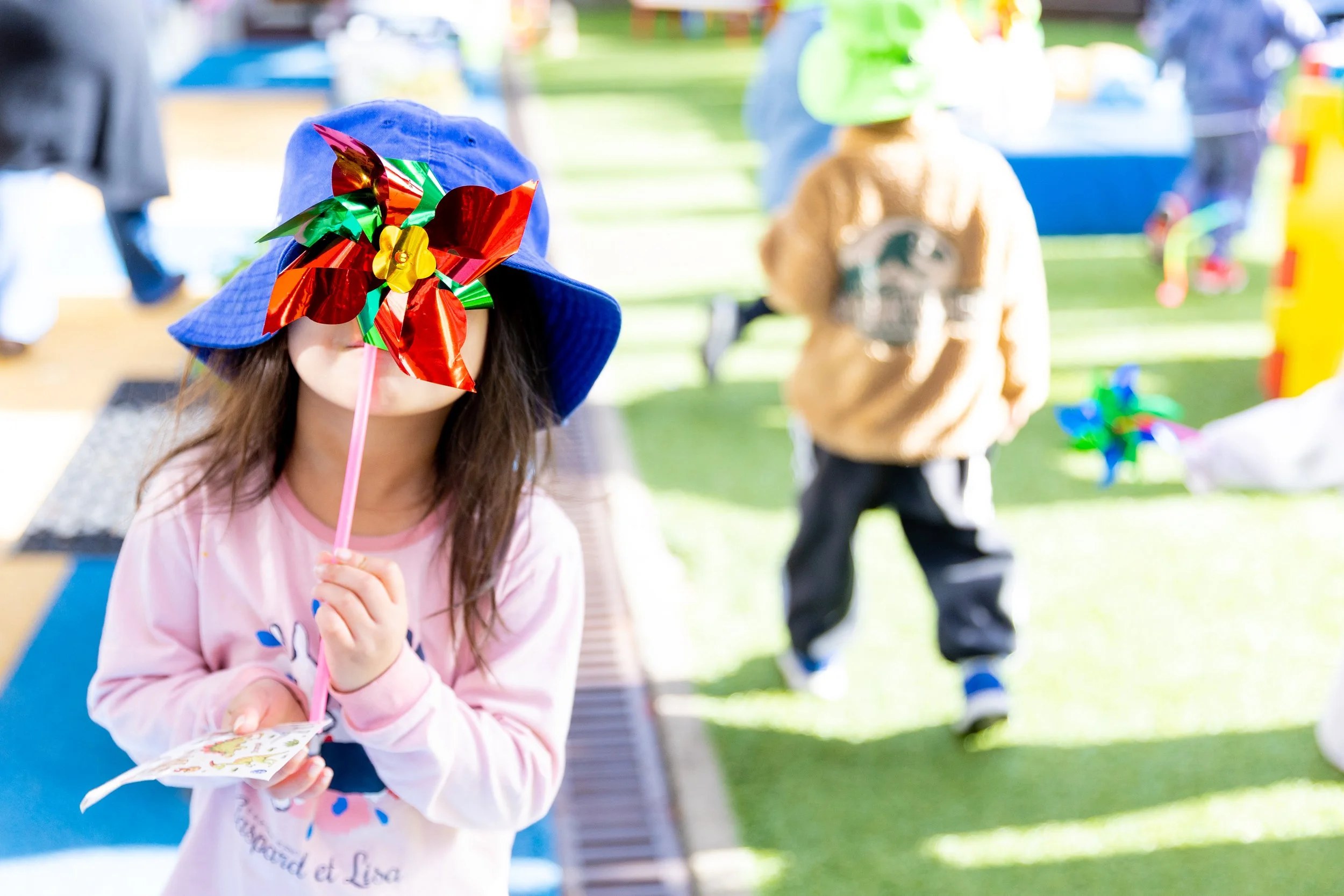 A young girl wearing a blue sunhat with a colorful pinwheel attached to it, holding some papers, at an outdoor event with other children and adults in the background.