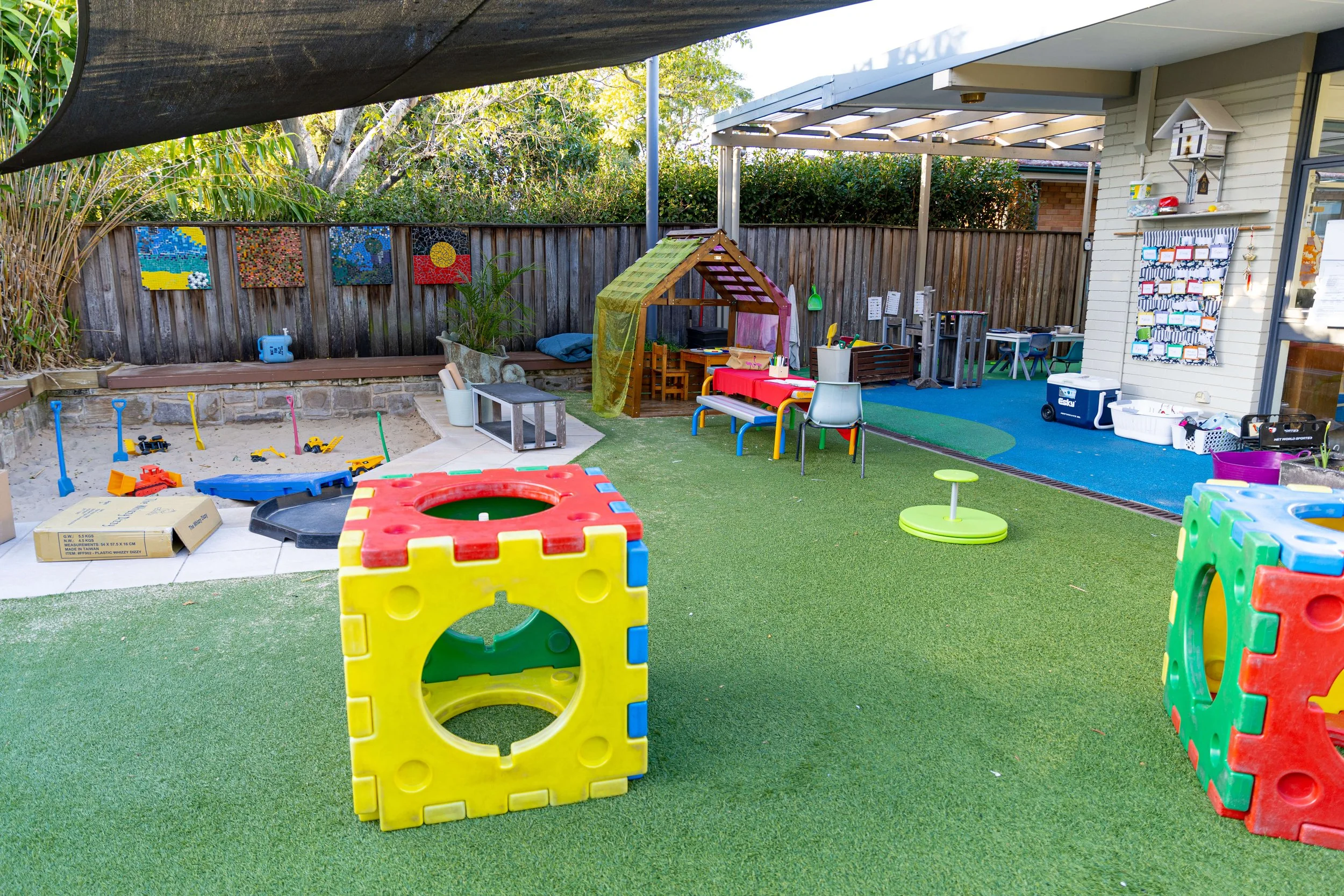 Children's outdoor play area with colorful plastic blocks, sandbox toys, a small playhouse, and various tables and chairs for activities, surrounded by a wooden fence and lush greenery.