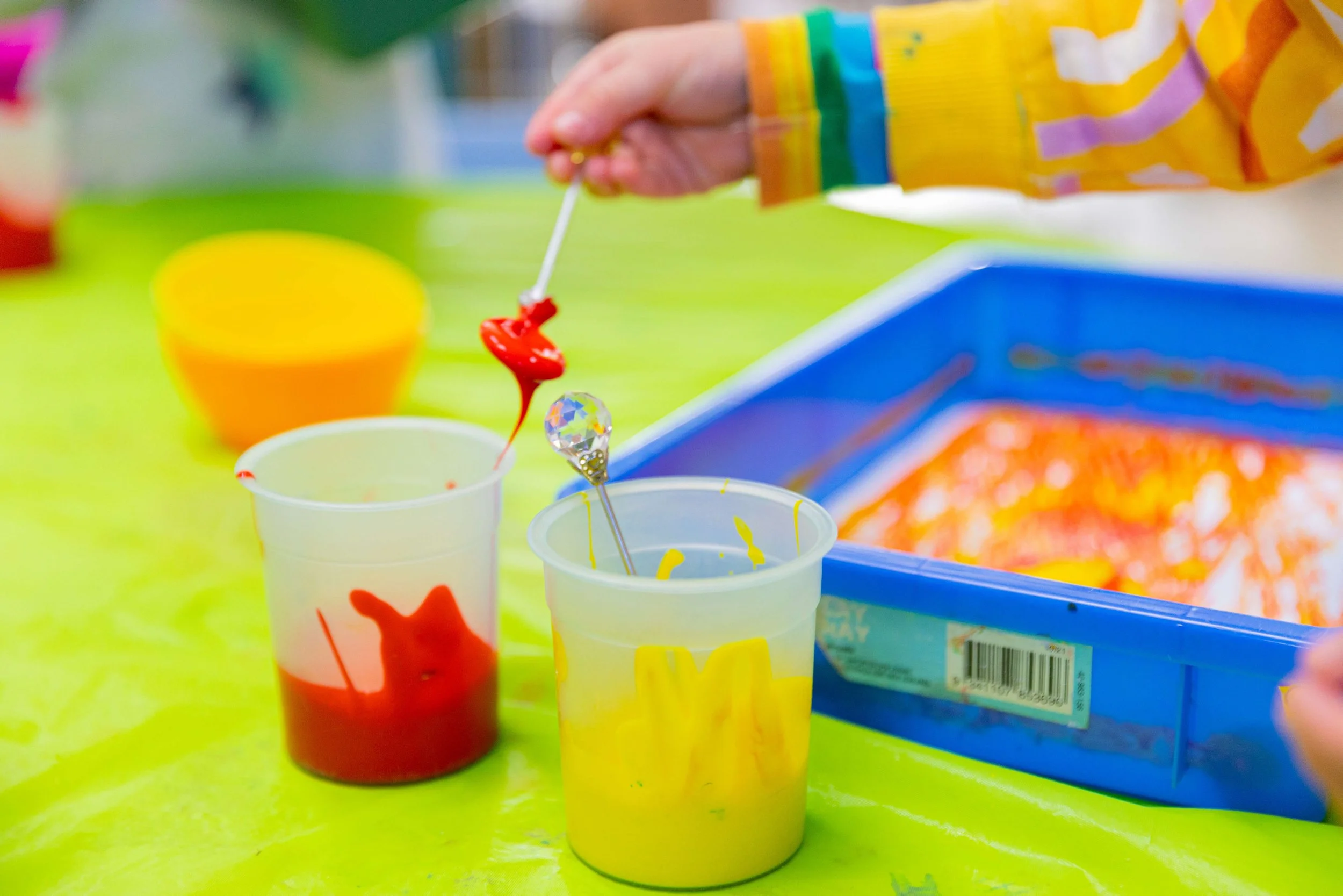 Child's hand stirring red and yellow paint in cups on a green table with art supplies