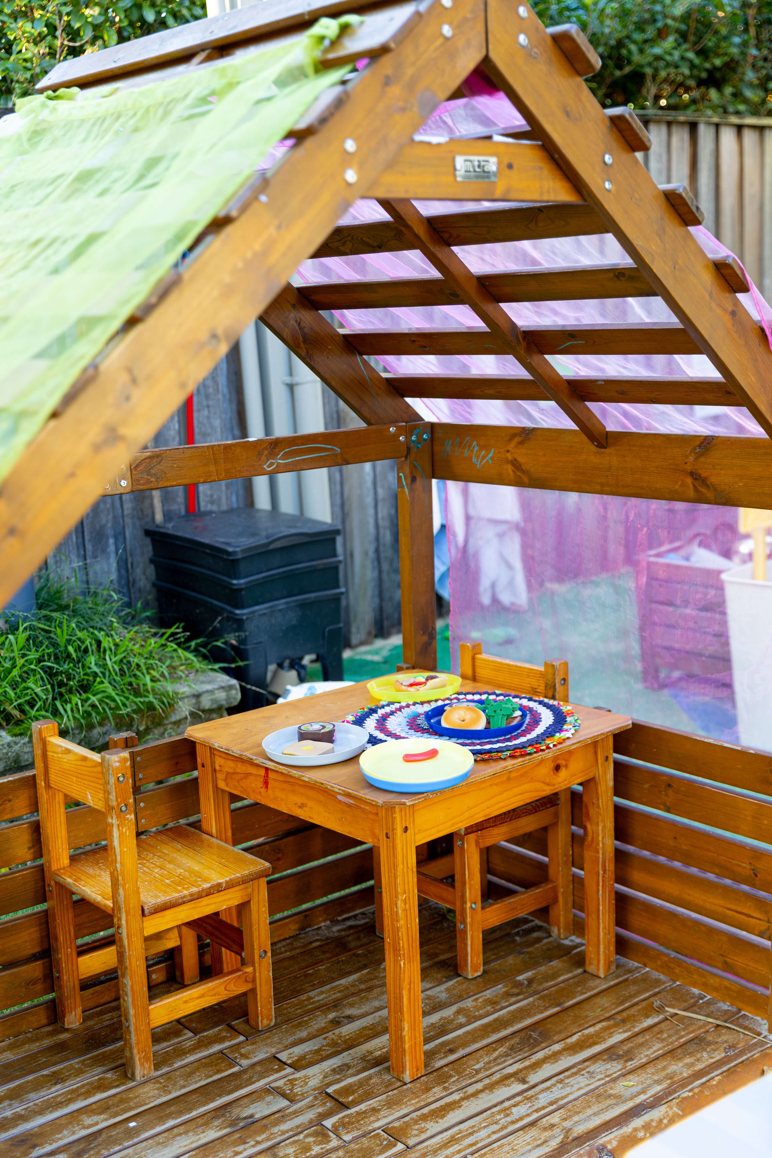 Children's outdoor playhouse with a small wooden table and chairs, decorated with toy food and play dishes, on a wooden deck with a roof and partial pink curtain.