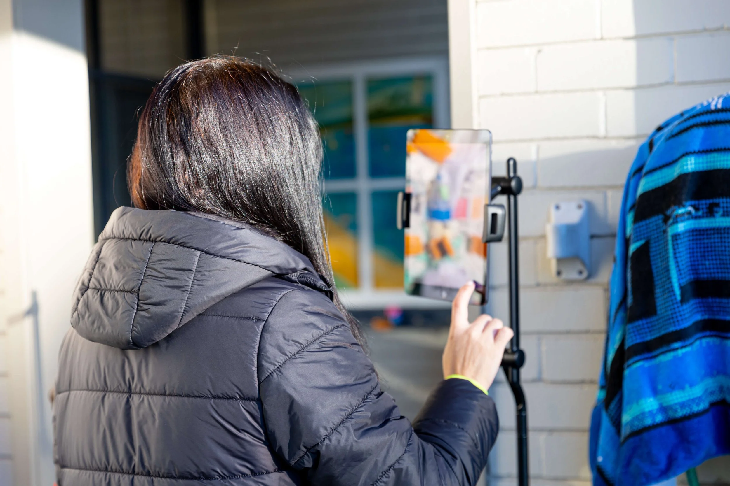 A person with dark hair wearing a gray puffer jacket takes a photo with a smartphone on a tripod outside near a white brick wall, with another person wearing a blue and black striped hoodie nearby.