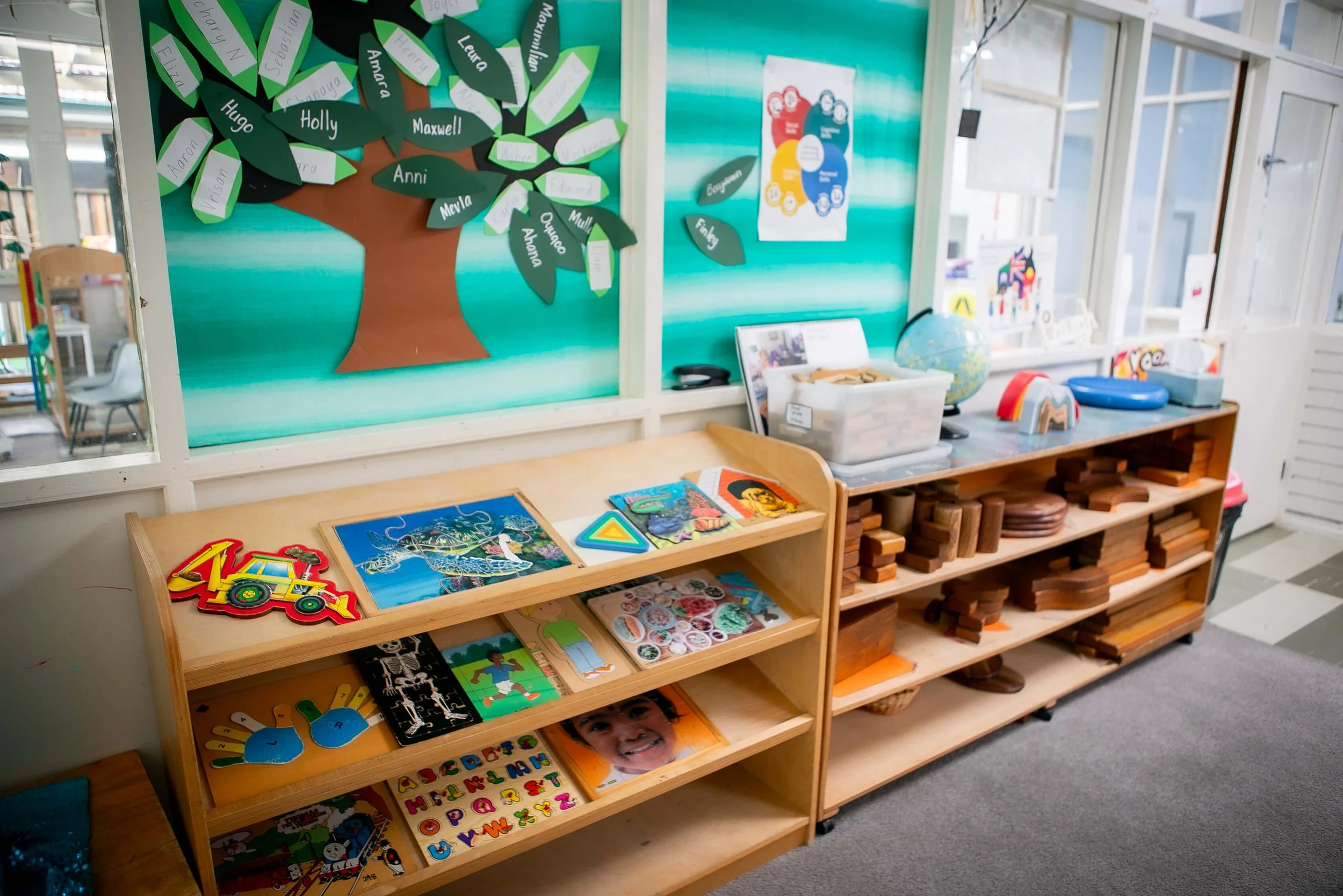 Child's classroom or play area with wooden shelves holding educational toys, puzzles, books, and wooden blocks. The background features a bulletin board decorated with a paper tree and paper leaves with children's names.