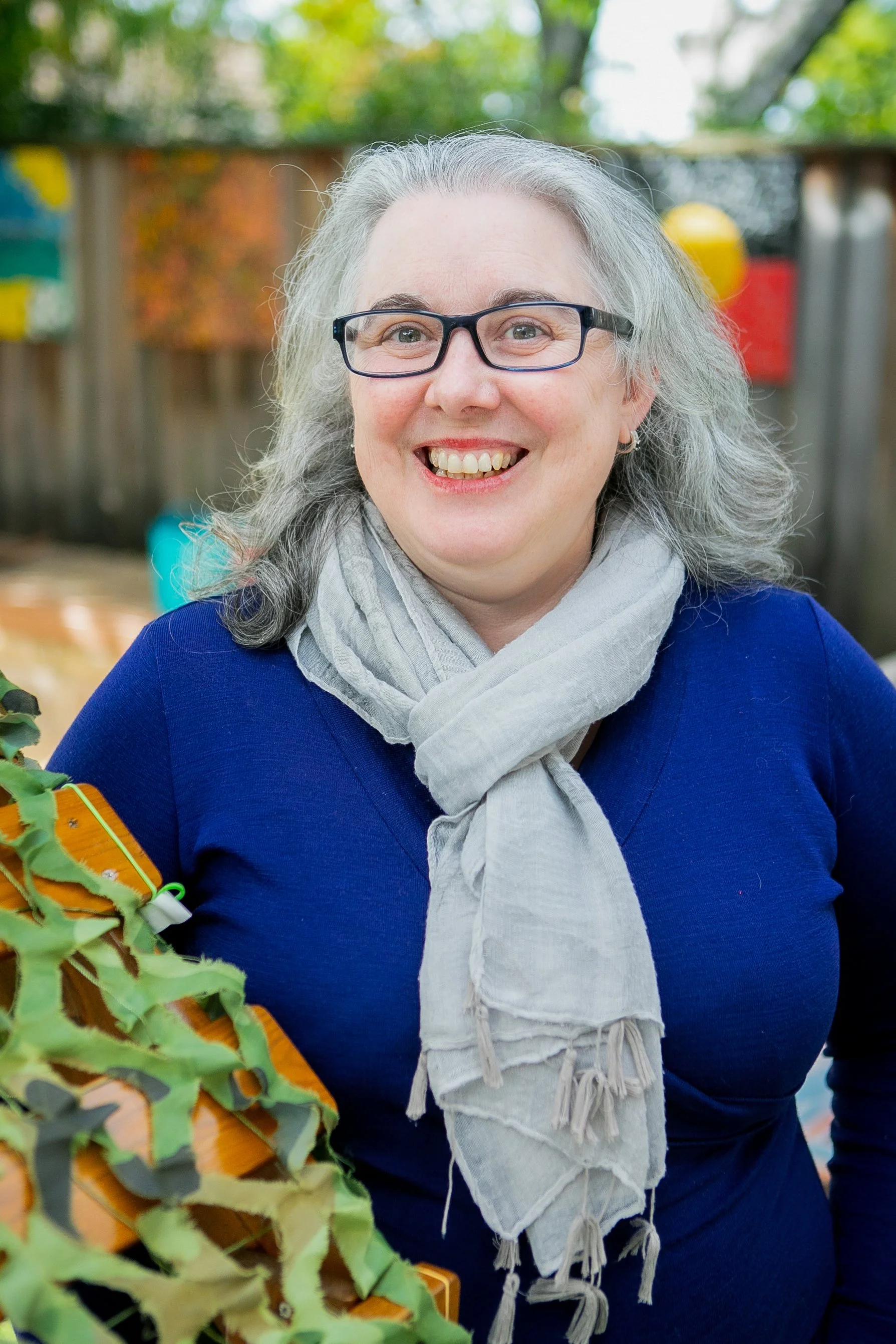 Woman with gray hair and glasses smiling outdoors, wearing a blue top and a light gray scarf, with a wooden fence and trees in the background.