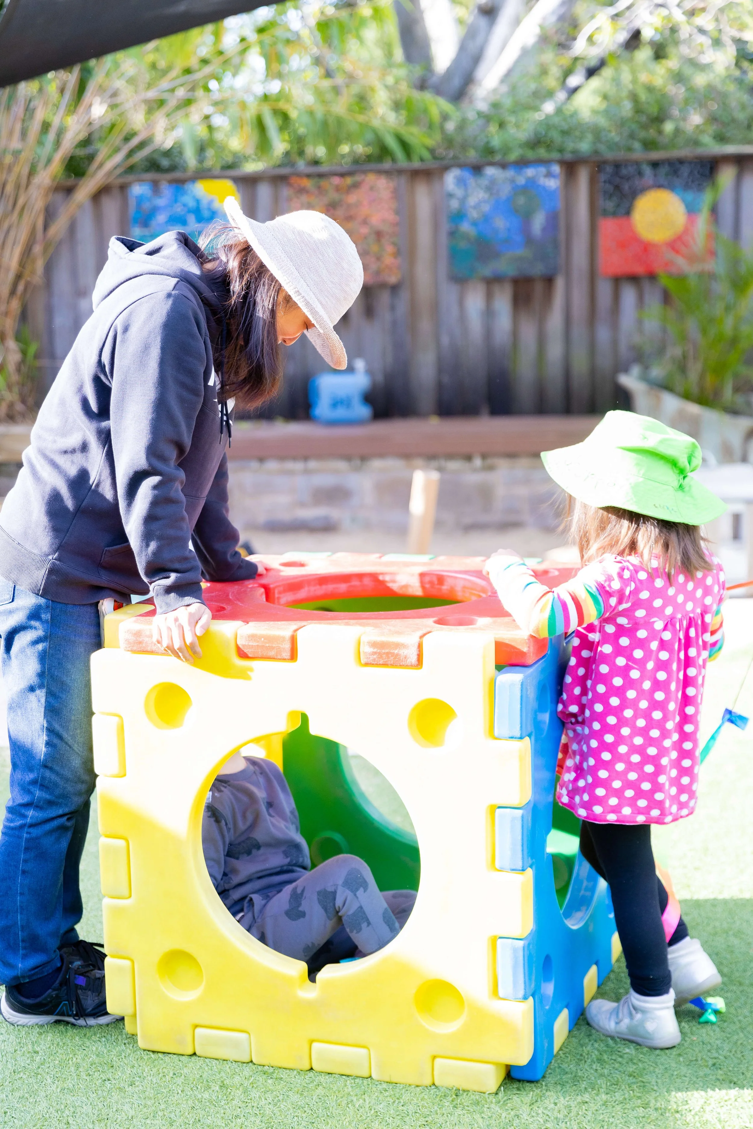 Two children and an adult playing on a colorful plastic cube-shaped outdoor play structure with round and square openings in a sunny backyard.